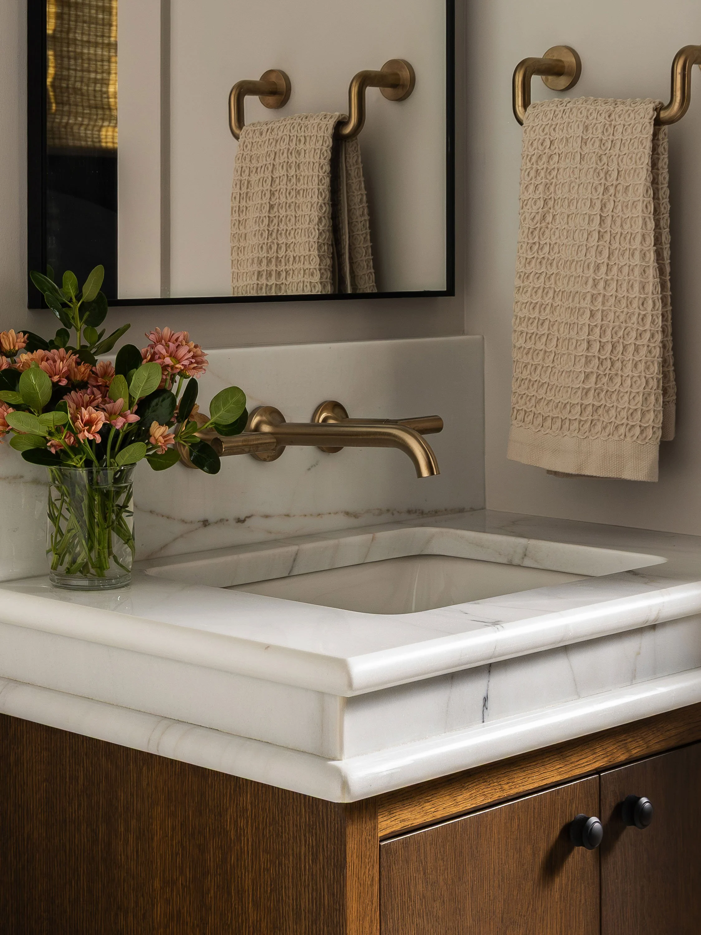 A bathroom sink with a marble countertop, a brass faucet, a mirror, a towel rack with a beige towel, and a vase with pink and orange flowers.