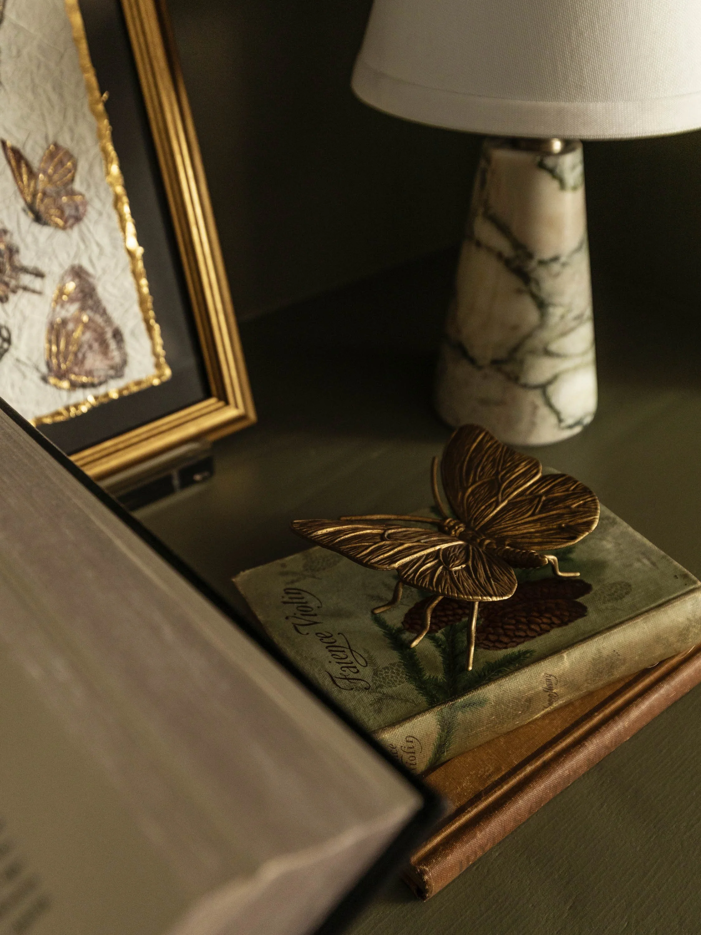 Close-up of a decorative butterfly figurine on a shelf next to a book with a marble lamp on top, with a framed butterfly artwork on the side.
