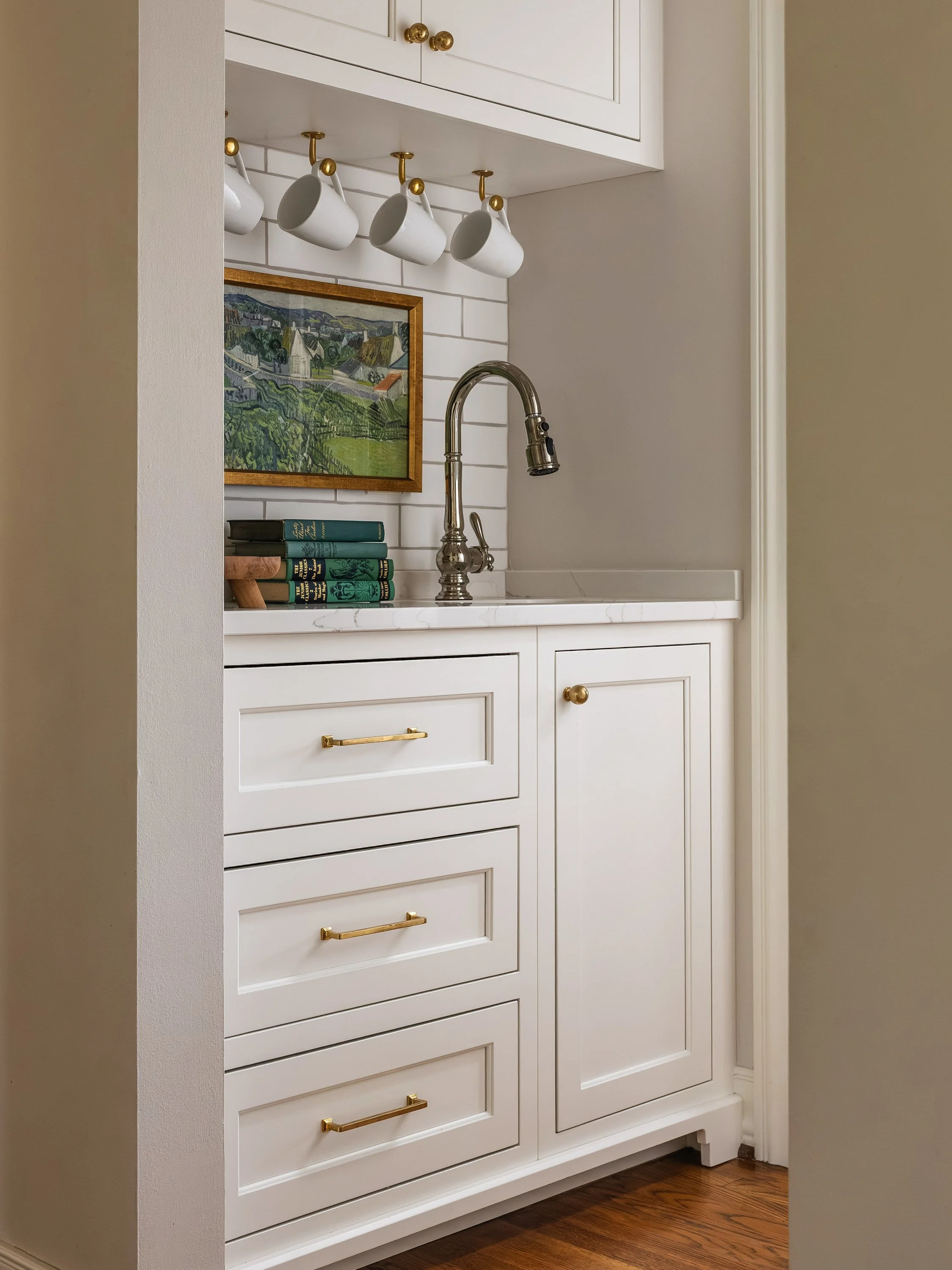 White kitchen cabinetry with drawers and upper cabinets, gold knobs and handles, a brass faucet, a framed landscape painting, and white coffee mugs hanging under the upper cabinet.