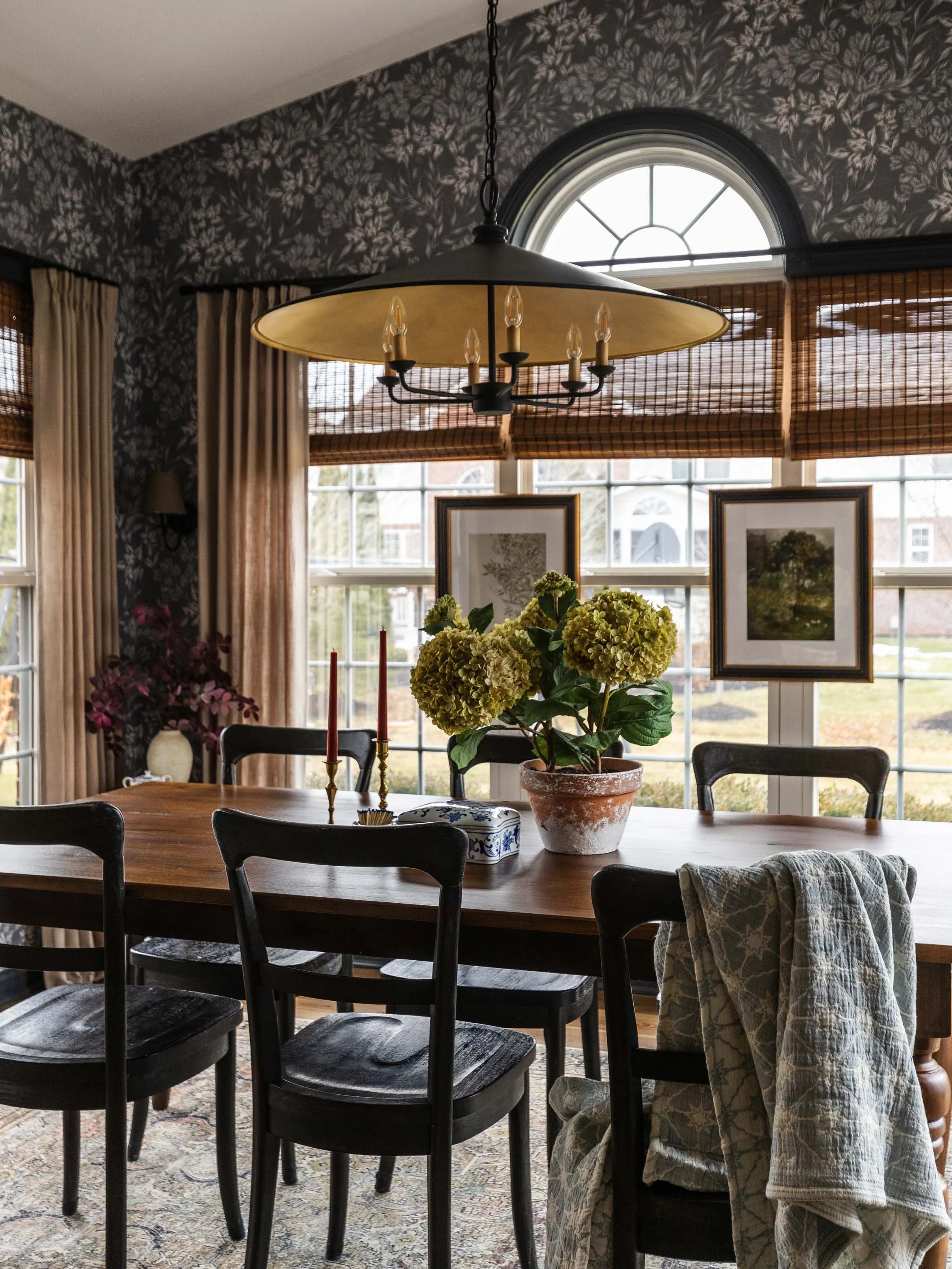 A dining room with a wooden table, black chairs, a potted hydrangea plant, a chandelier hanging from the ceiling, framed artwork on the window, and floral wallpaper with curtains, next to windows with bamboo blinds.