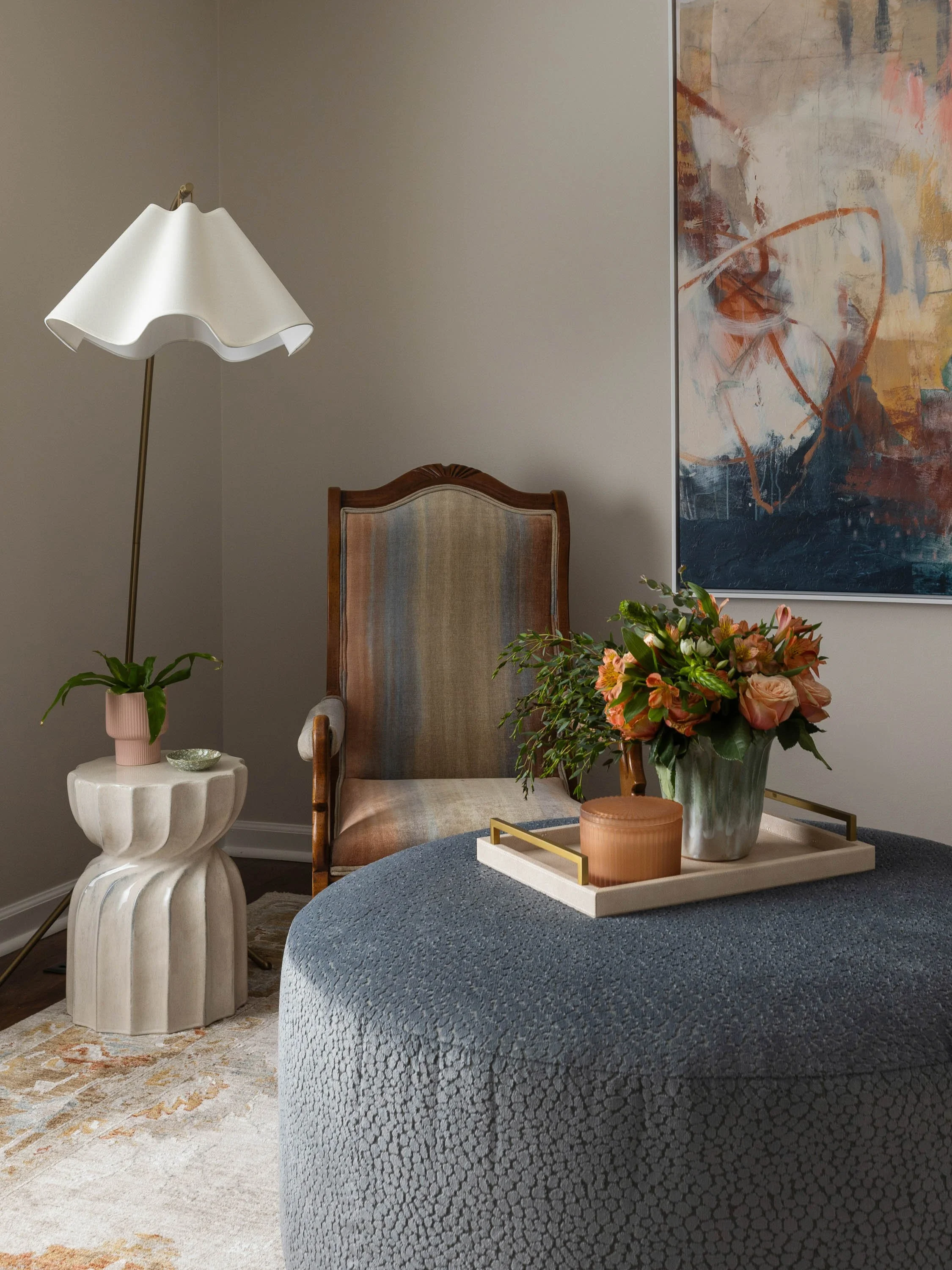 A living room corner with a tall white sculptural side table, a wooden armchair, a side table with a flower arrangement, a textured gray ottoman, a white tray with decorative items, a modern standing lamp, abstract wall art, and a rug with a neutral 