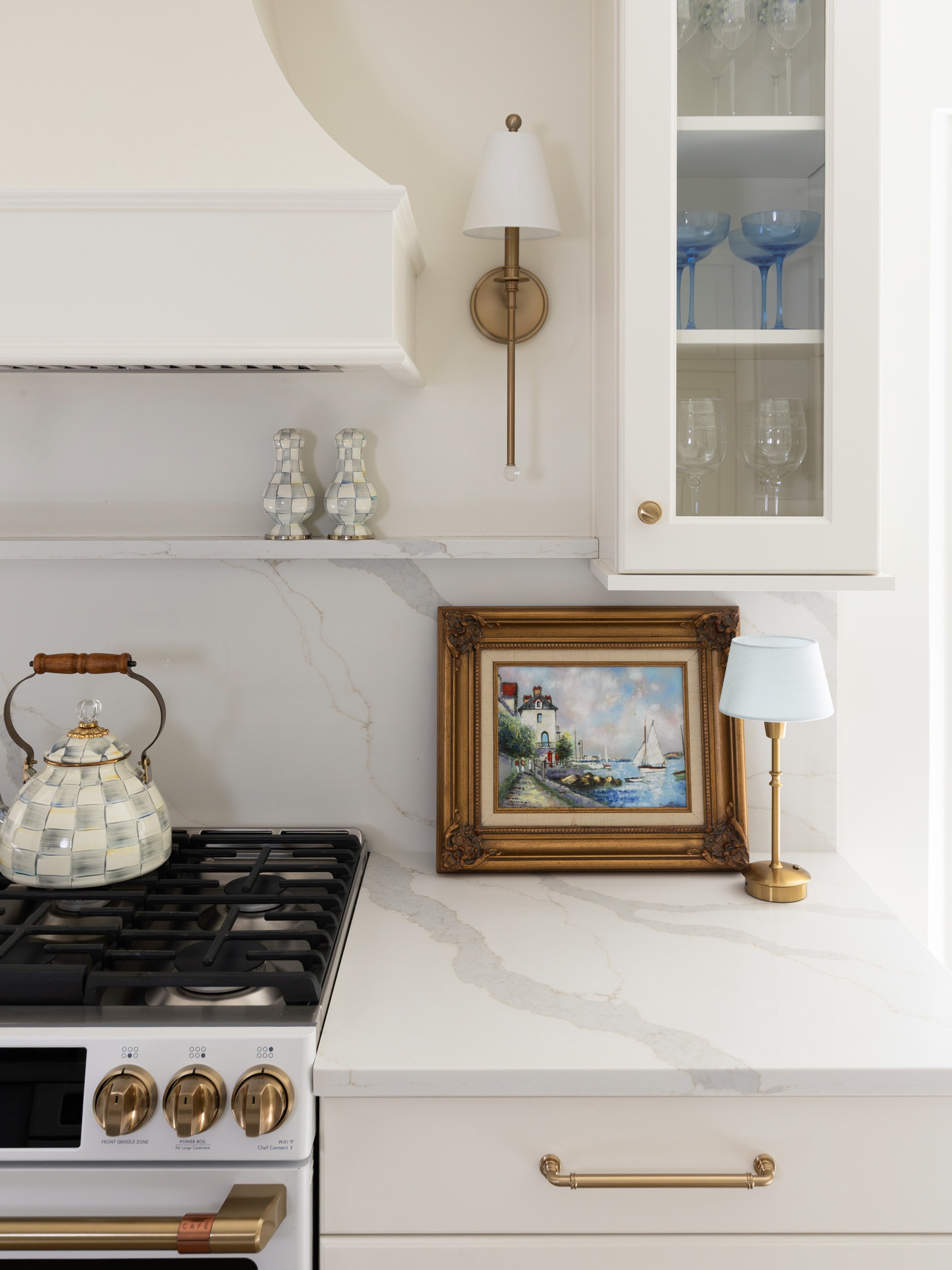 A kitchen countertop with a gold and white teapot, a framed painting of a lighthouse scene, a gold lamp, and a white cabinet with glass doors containing glasses and bowls. The stove has gold knobs.