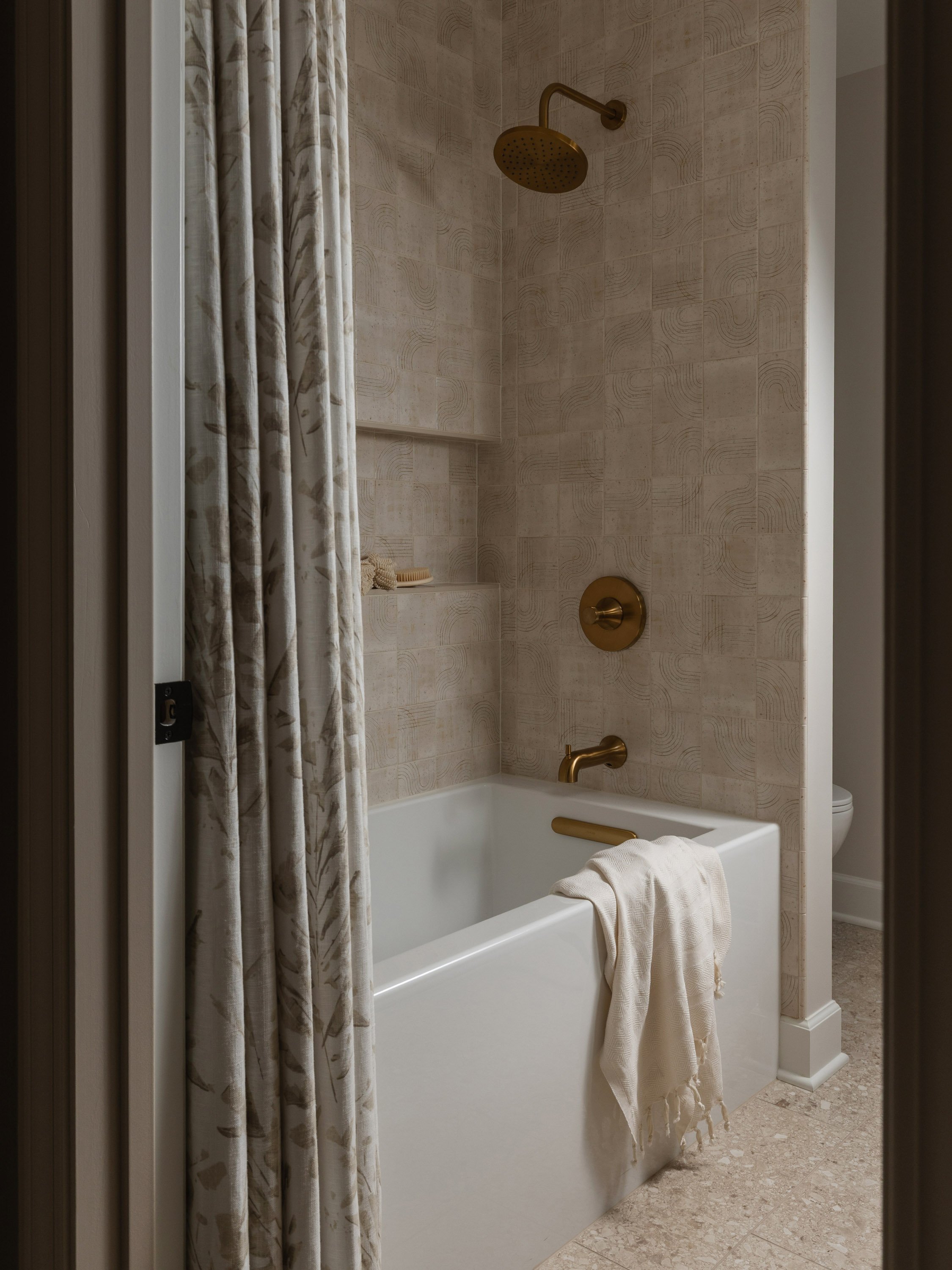 A bathroom shower with a beige tiled wall, brass showerhead, and a white bathtub. A beige towel hangs on the edge, and a shower curtain with a subtle leaf pattern is partially closed. A small shelf holds a brush and loofah.
