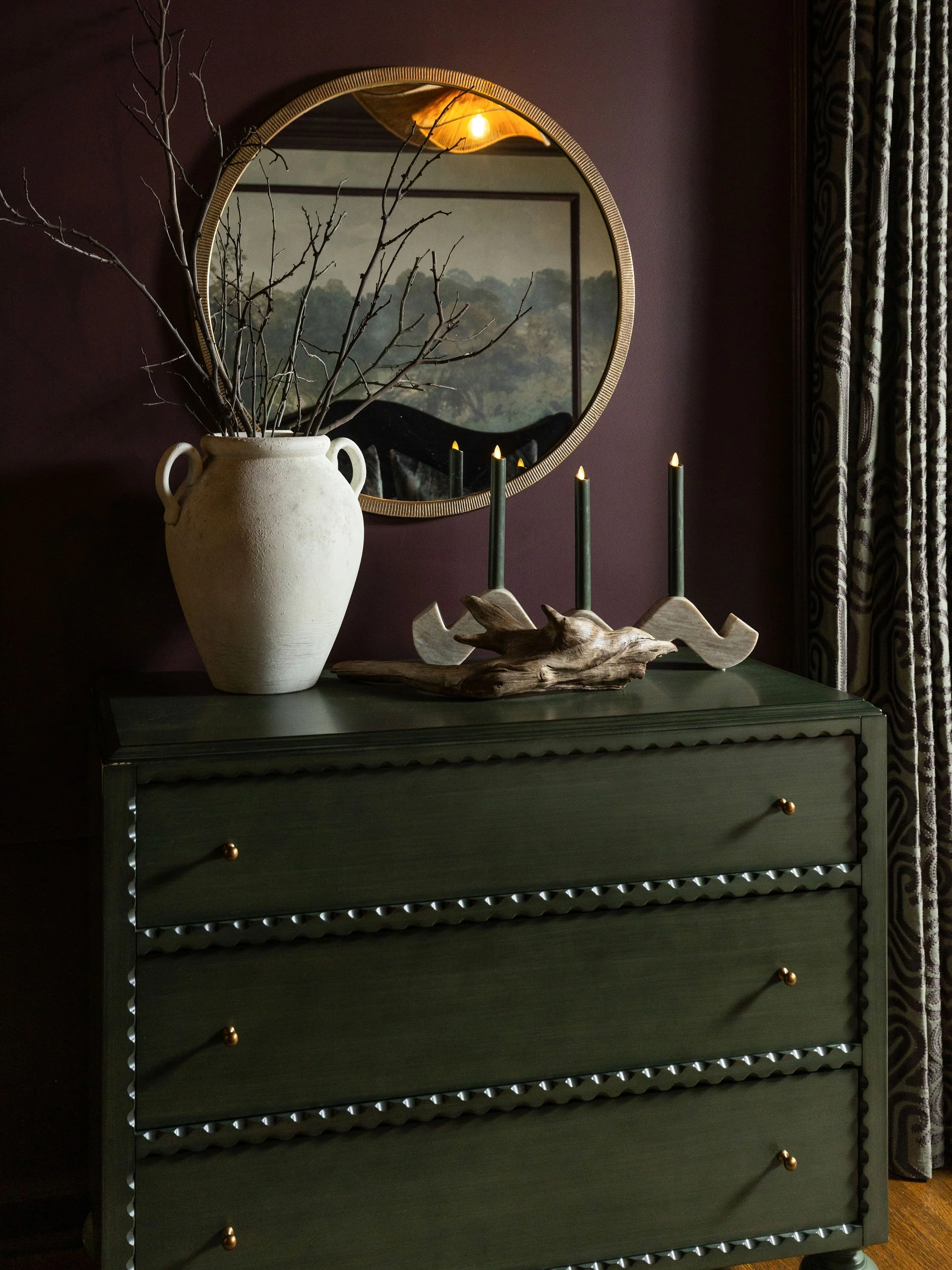 A dark green dresser with gold knobs, decorated with a large ceramic vase holding bare branches, a wooden branch used as candle holders with three lit candles, and a mirror above reflecting a painting and a light fixture. There are curtains with a bl