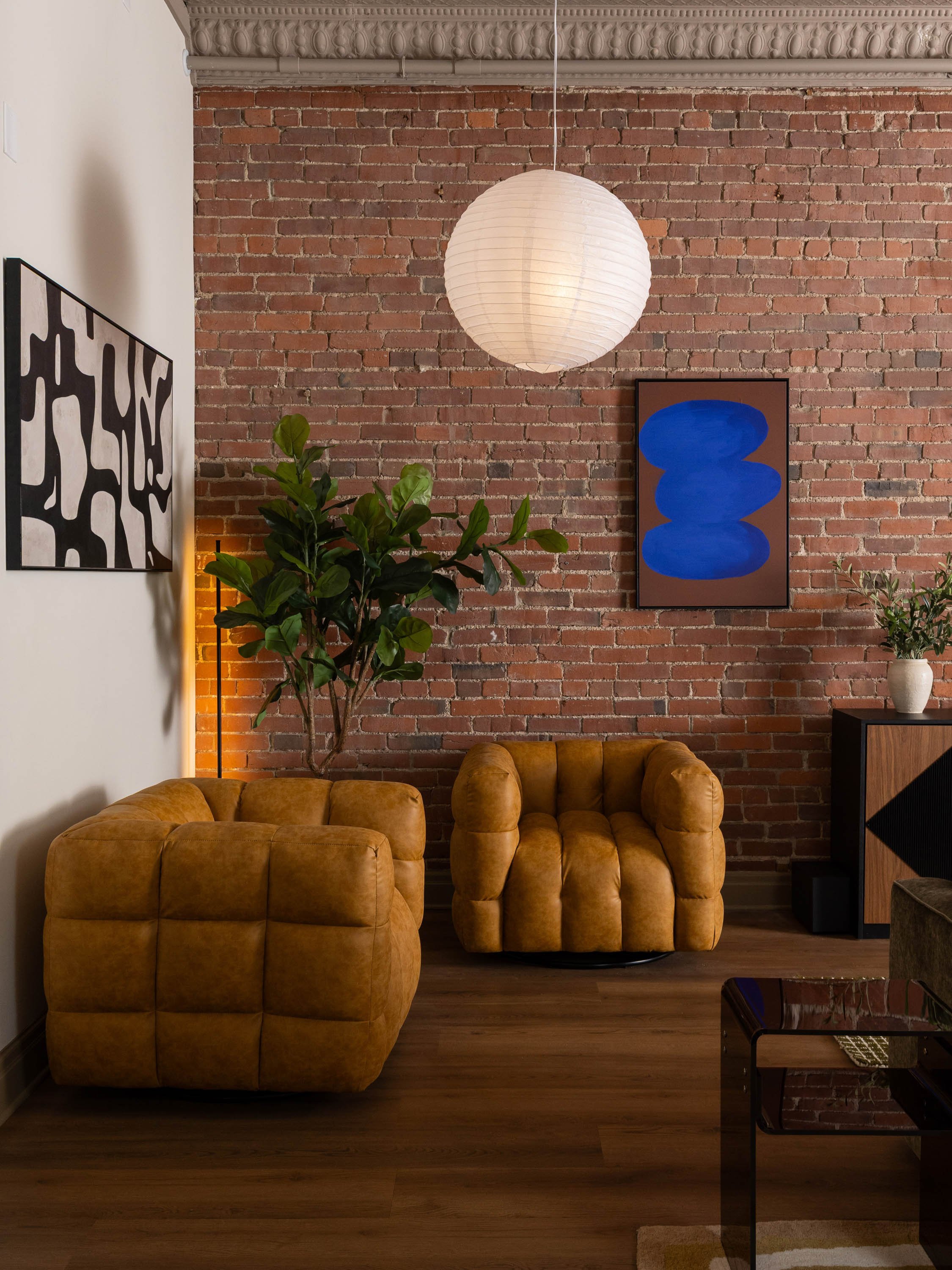 Living room with a brick wall, two mustard-yellow tufted armchairs, a potted green plant, framed abstract artwork with black and white pattern, a white paper lantern hanging from the ceiling, a black side table with a glass top, and a wooden cabinet 