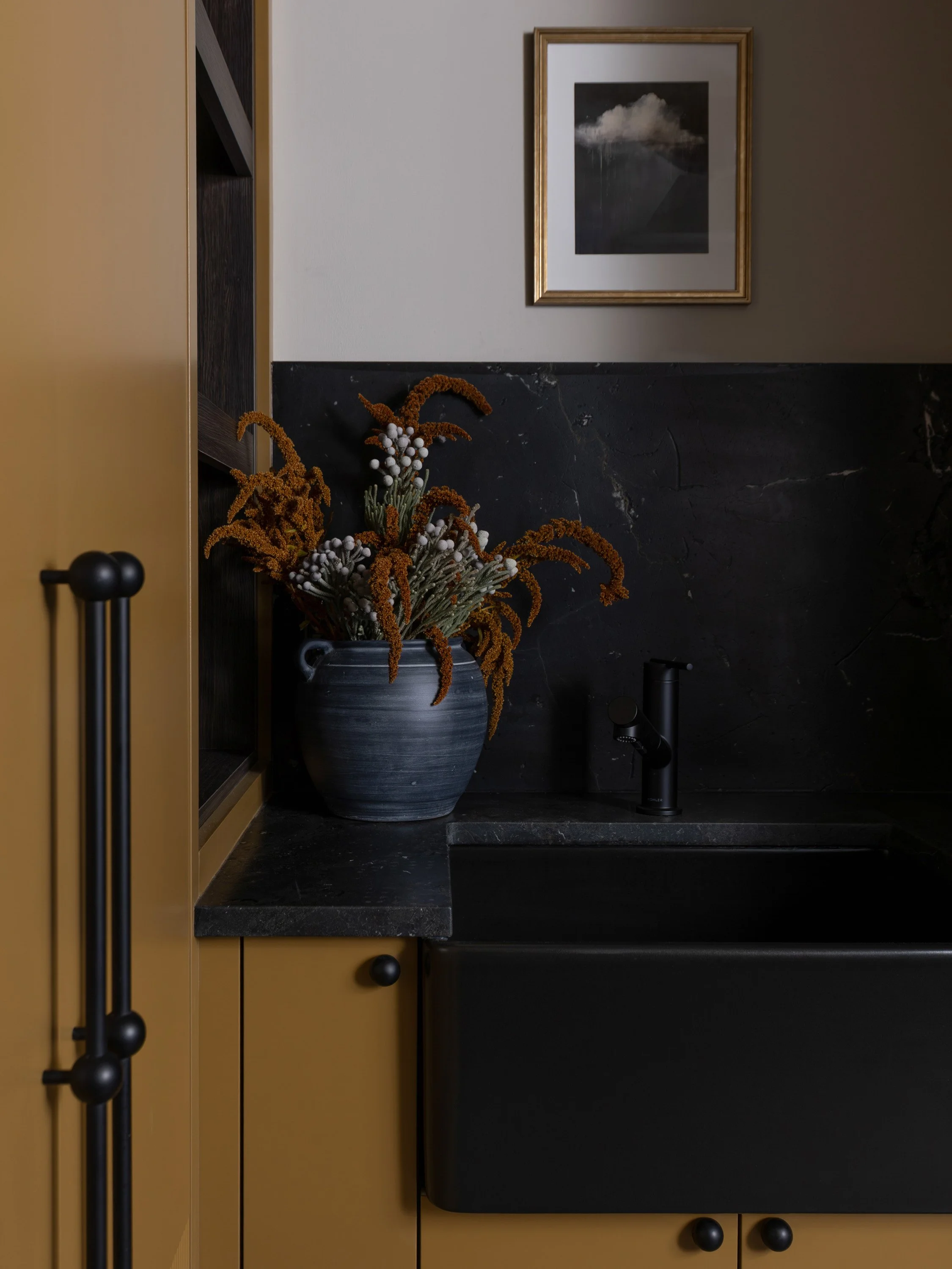 Interior view of a modern kitchen corner with a black sink, a decorative vase with dried orange and white flowers, and a framed black and white cloud photograph on the wall.