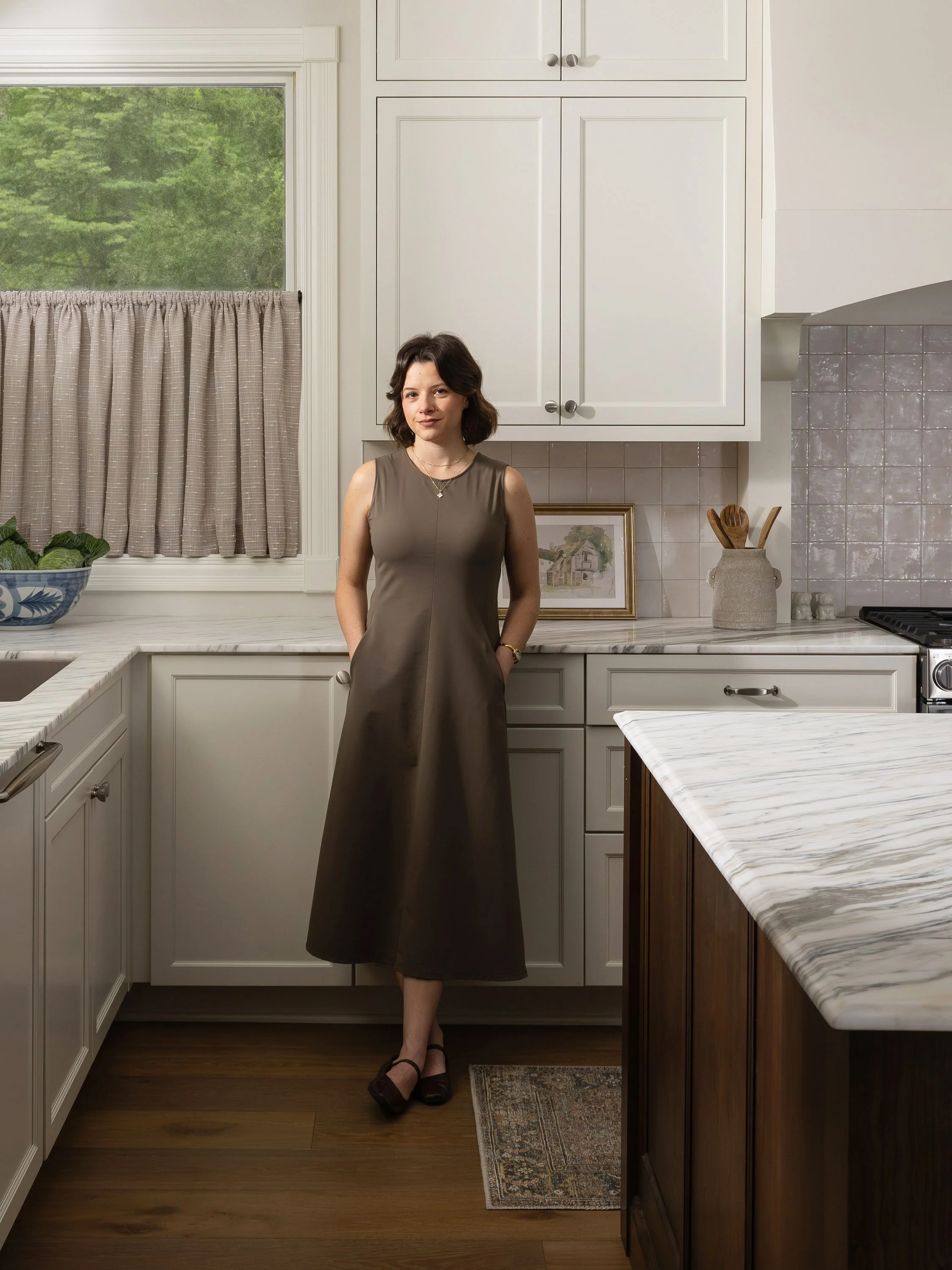 A woman in a brown dress standing in a kitchen between white cabinetry, with a window decorated with beige curtains behind her, and a marble-topped island in front of her.