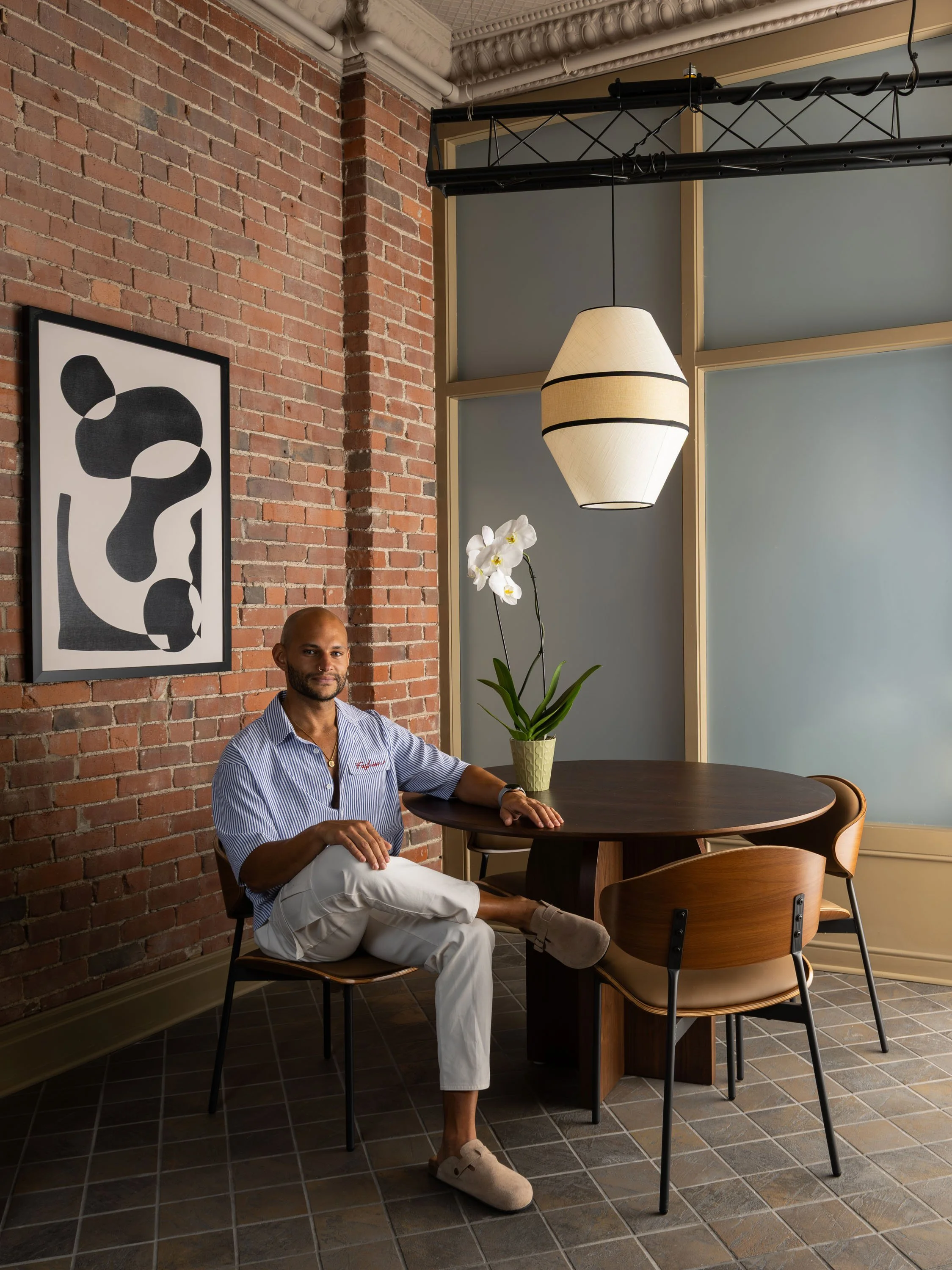 A man with dark skin, bald head, and beard is sitting at a round wooden table in a room with a brick wall. He is wearing a light blue and white striped shirt, white pants, and tan slippers. On the table, there is a potted white orchid flower. Behind 
