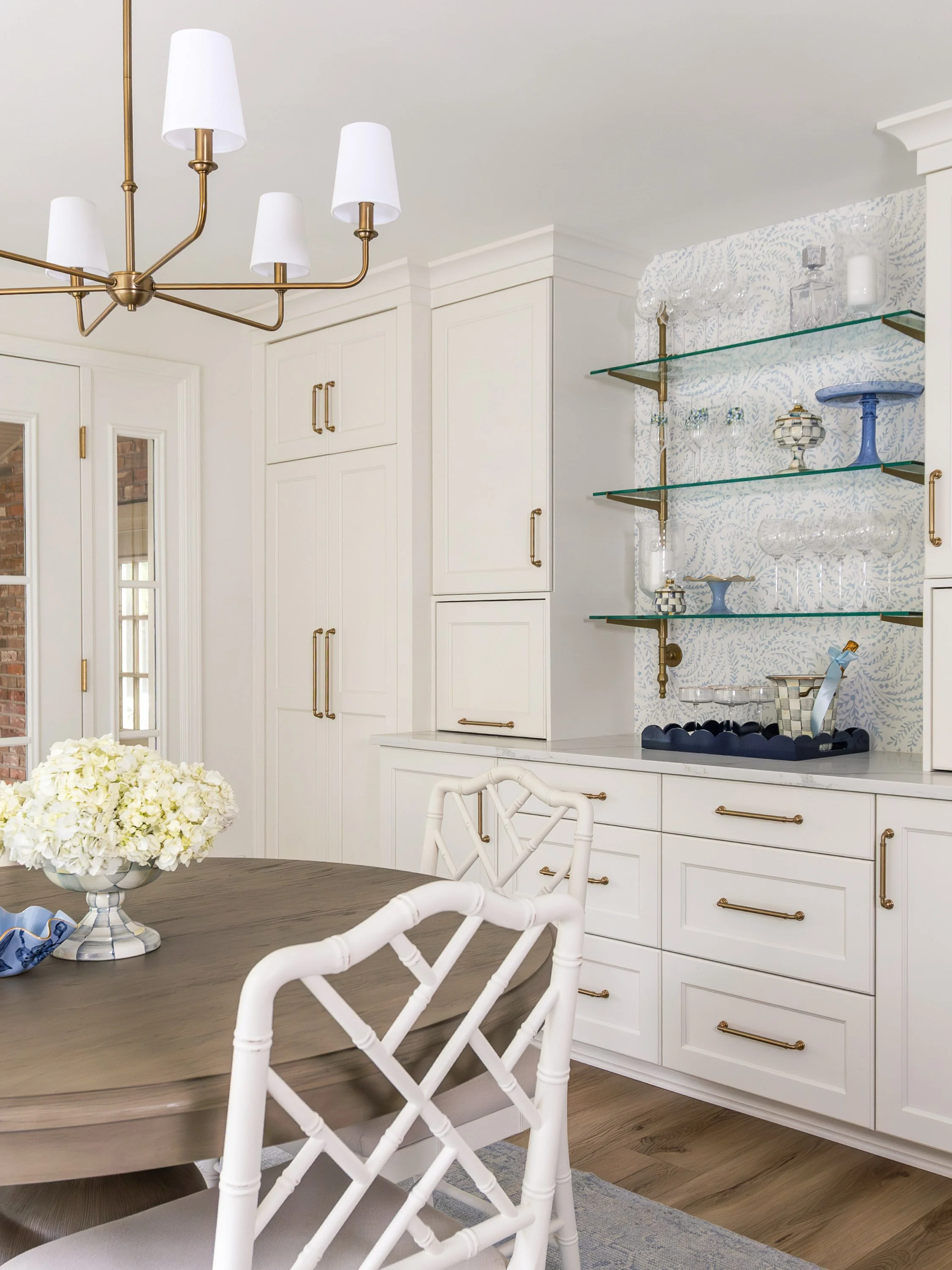 A modern dining room with a wooden table, white chairs, a floral centerpiece, white built-in cabinets, and glass shelves with decorative glassware and vases. A gold chandelier hangs from the ceiling.
