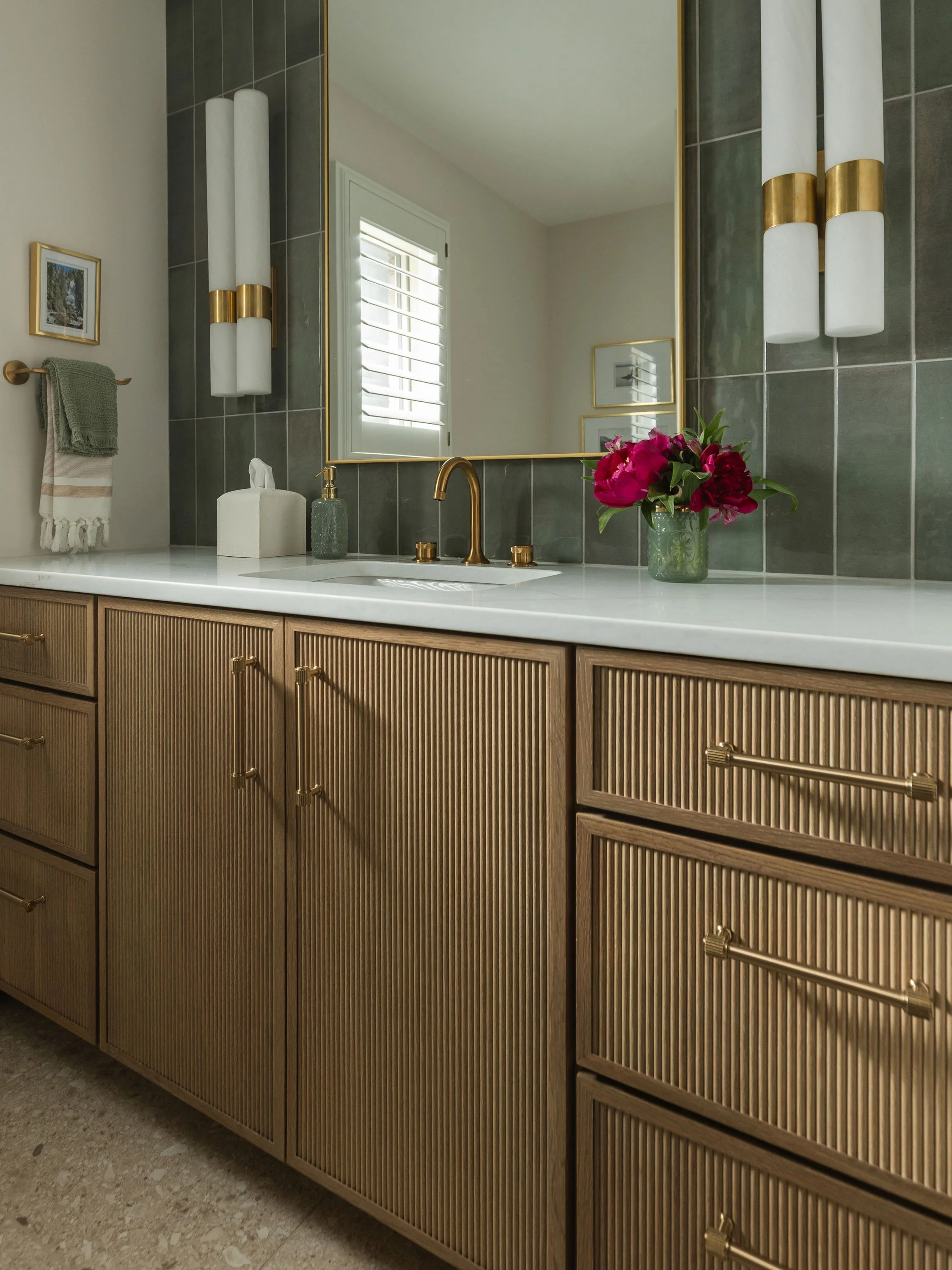 Bathroom vanity with wooden cabinets, a white marble countertop, a gold faucet, a glass soap dispenser, a tissue box, a flower vase with pink flowers, a mirror, wall-mounted light fixtures, a towel on a rack, and framed pictures on the wall.