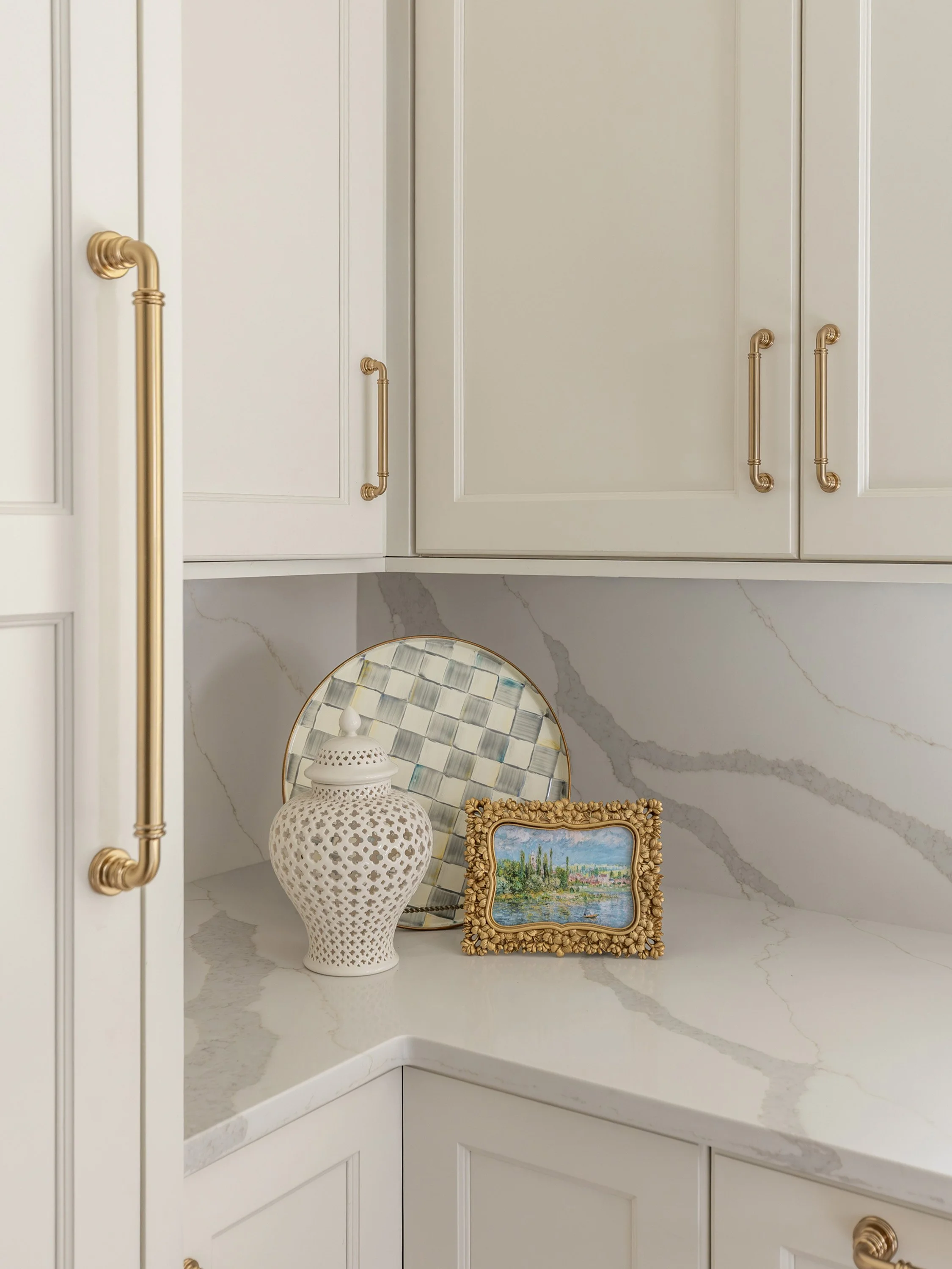 White kitchen cabinets with gold handles, a marble countertop, and decorative items including a framed landscape painting, a ceramic vase, and a woven tray.