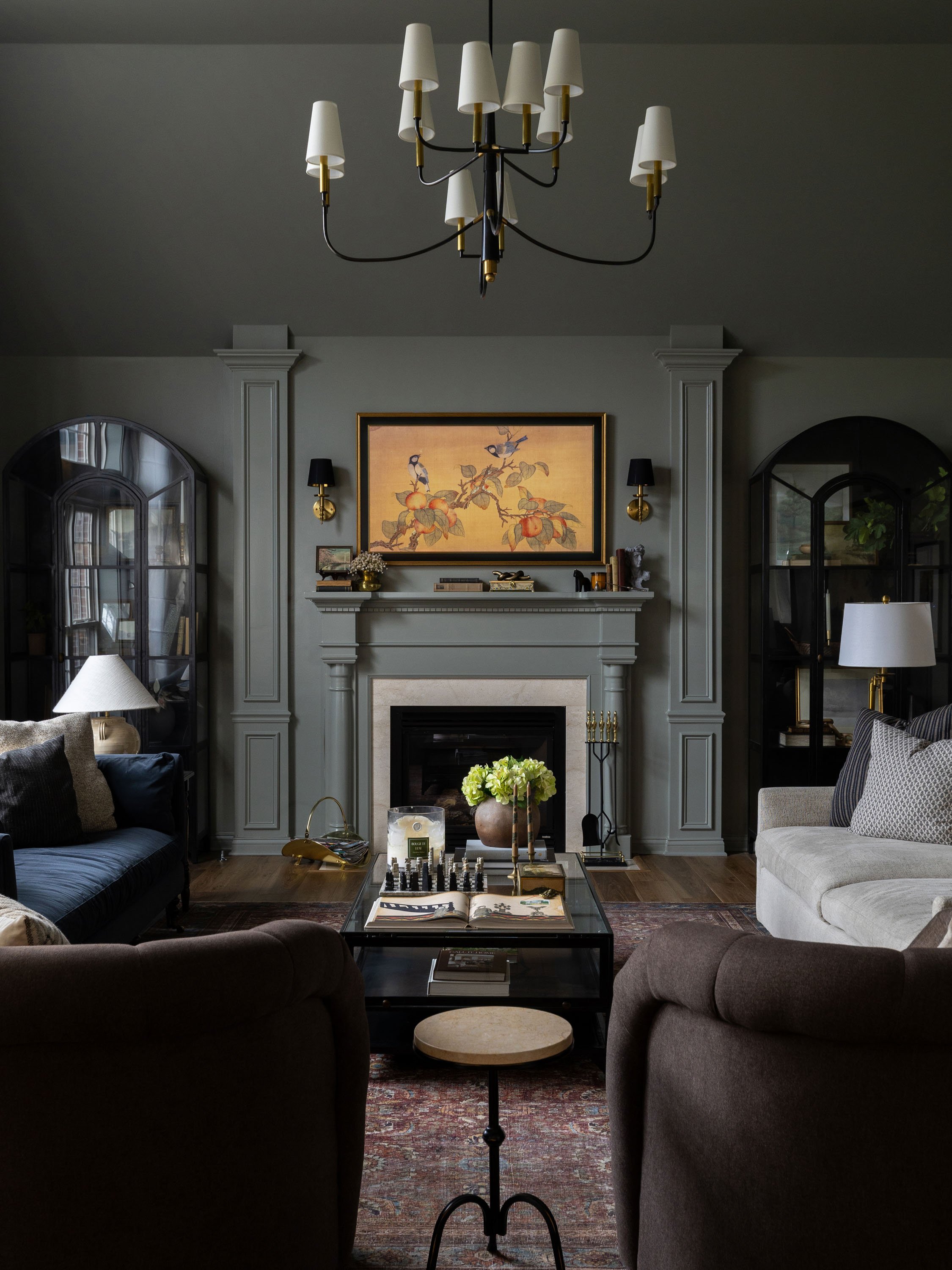 A cozy living room with gray walls and a fireplace, decorated with artwork, candles, and books. Two sofas face each other, a glass coffee table in the center holds a flower arrangement, books, and a chess set. There are two black cabinets flanking a 