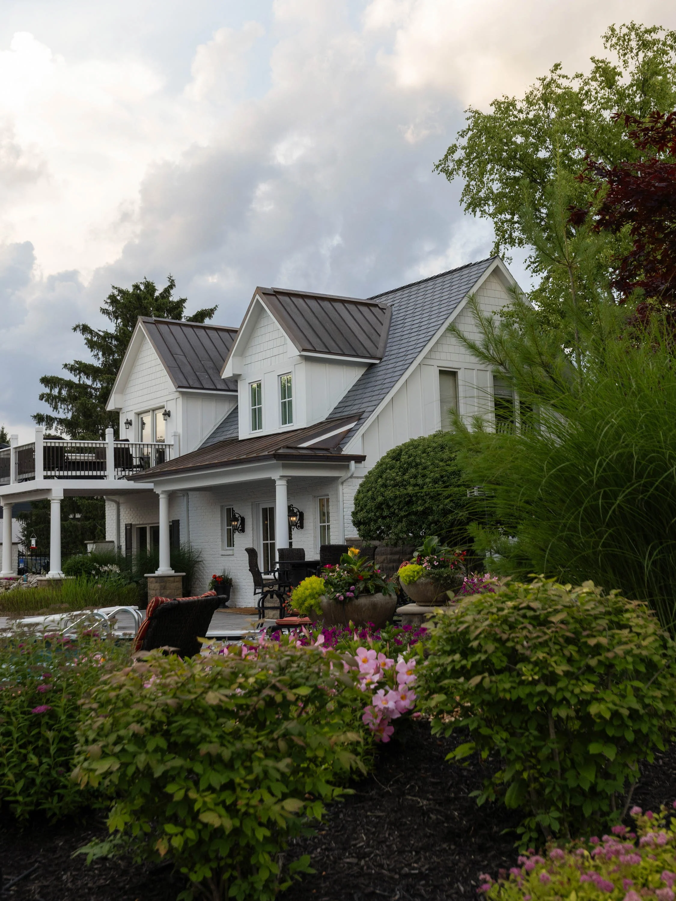 A white house with a deck and patio chairs surrounded by colorful flowers and greenery under a cloudy sky.