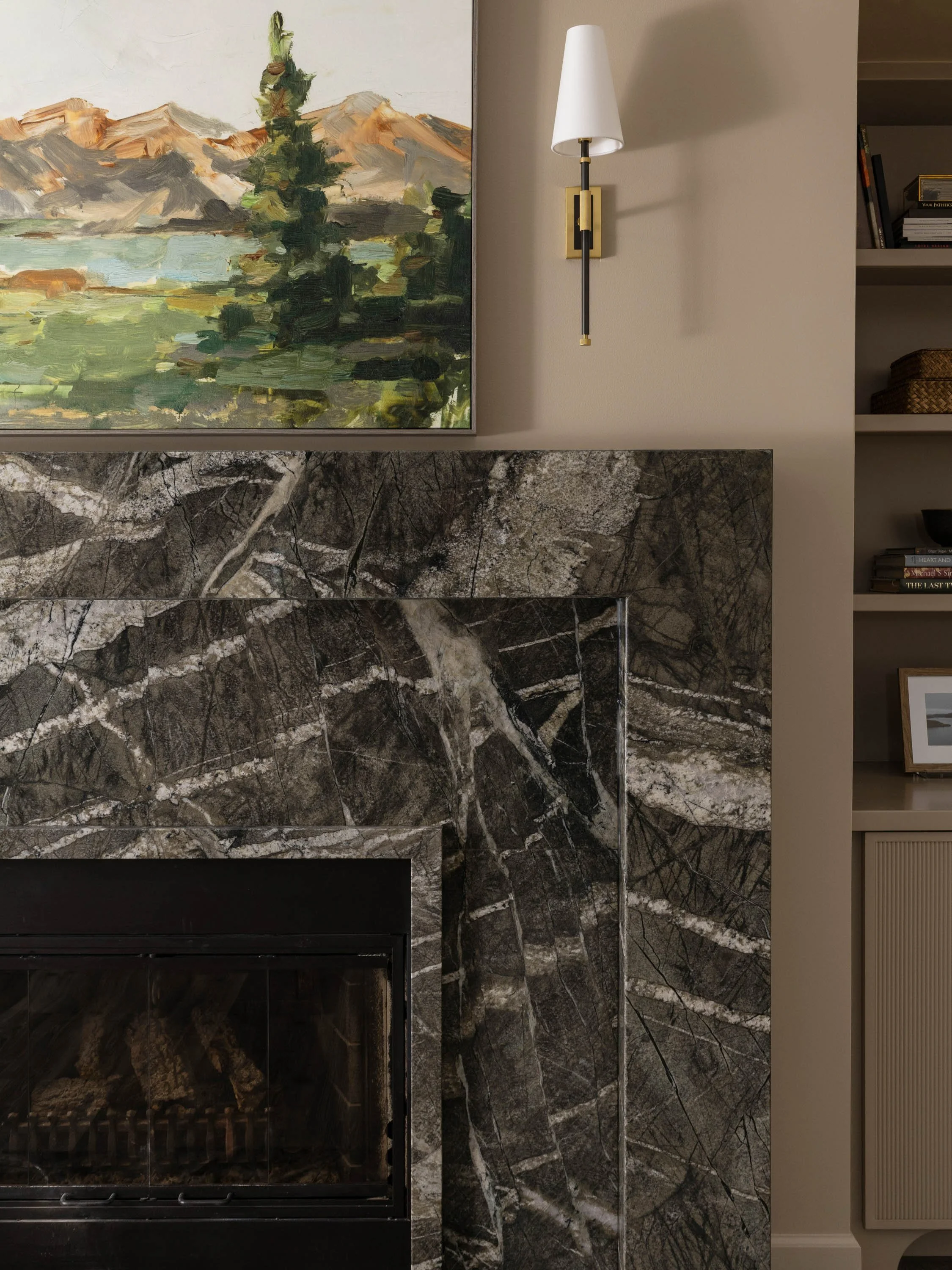Close-up of a marble fireplace with a granite surround, wooden bookshelf with books, an abstract landscape painting, and a wall-mounted modern wall lamp in a living room.