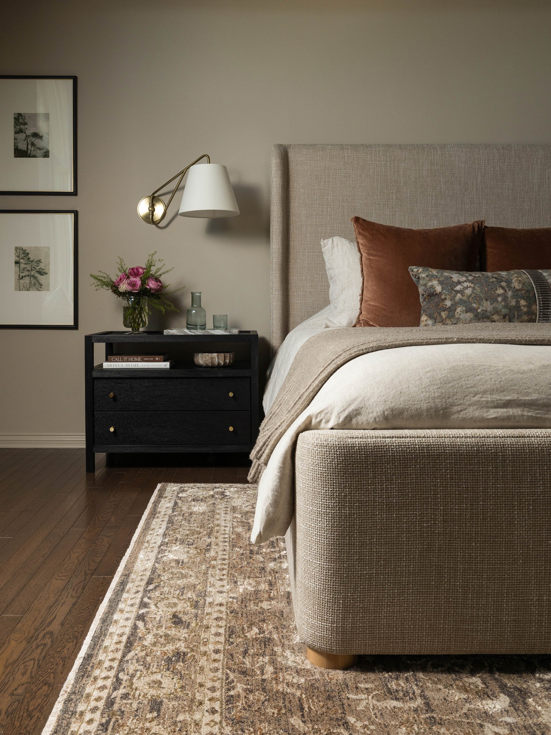 A bedroom corner with a beige upholstered bed, pillows, a black nightstand with a vase of pink flowers, a glass bottle, and picture frames on the wall.
