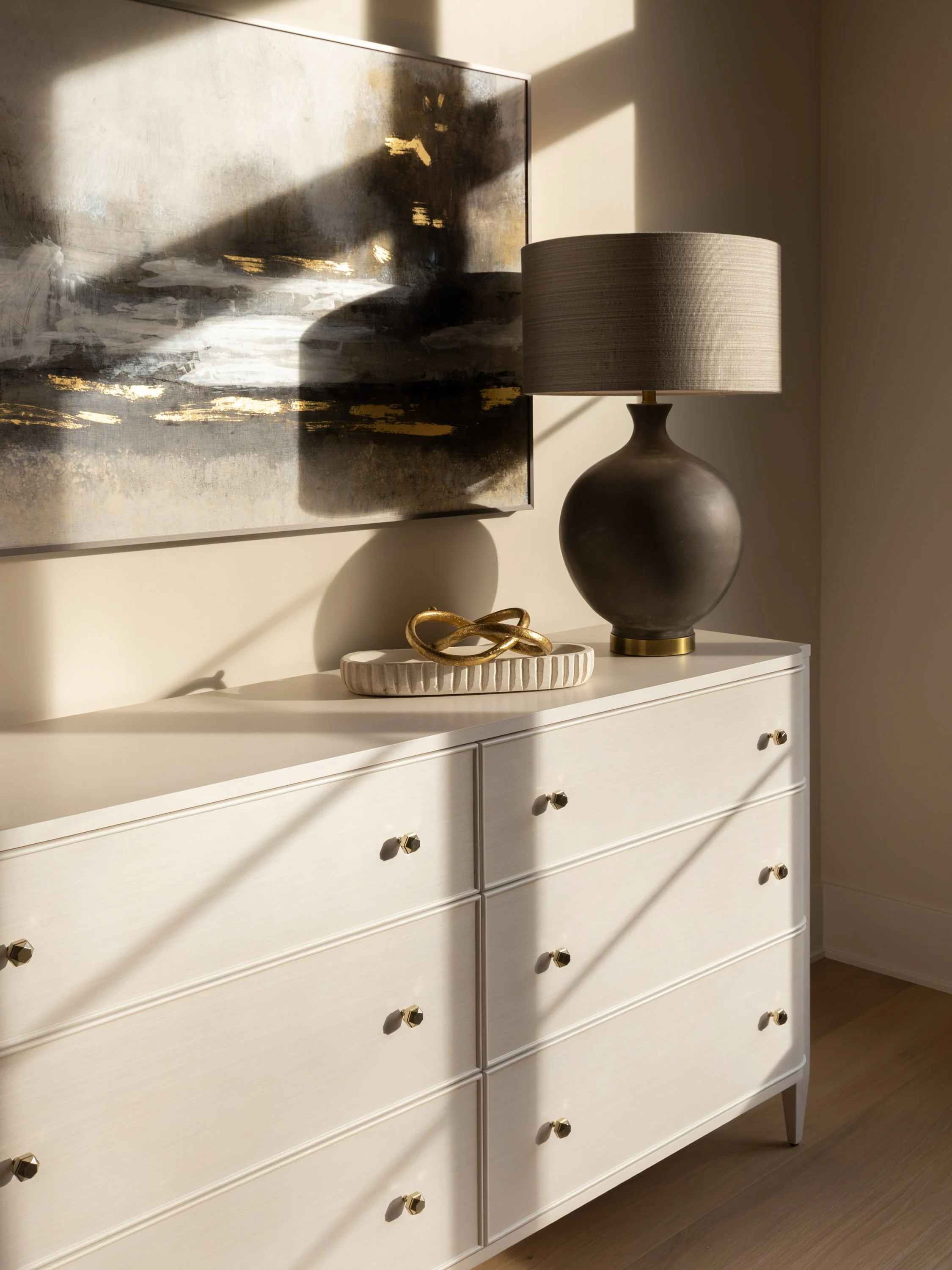 A white dresser with rounded edges and six drawers, a large dark ceramic table lamp with a beige fabric shade, a decorative gold orb sculpture on a white ceramic tray, and a modern abstract painting with black, white, and gold brushstrokes on the wal
