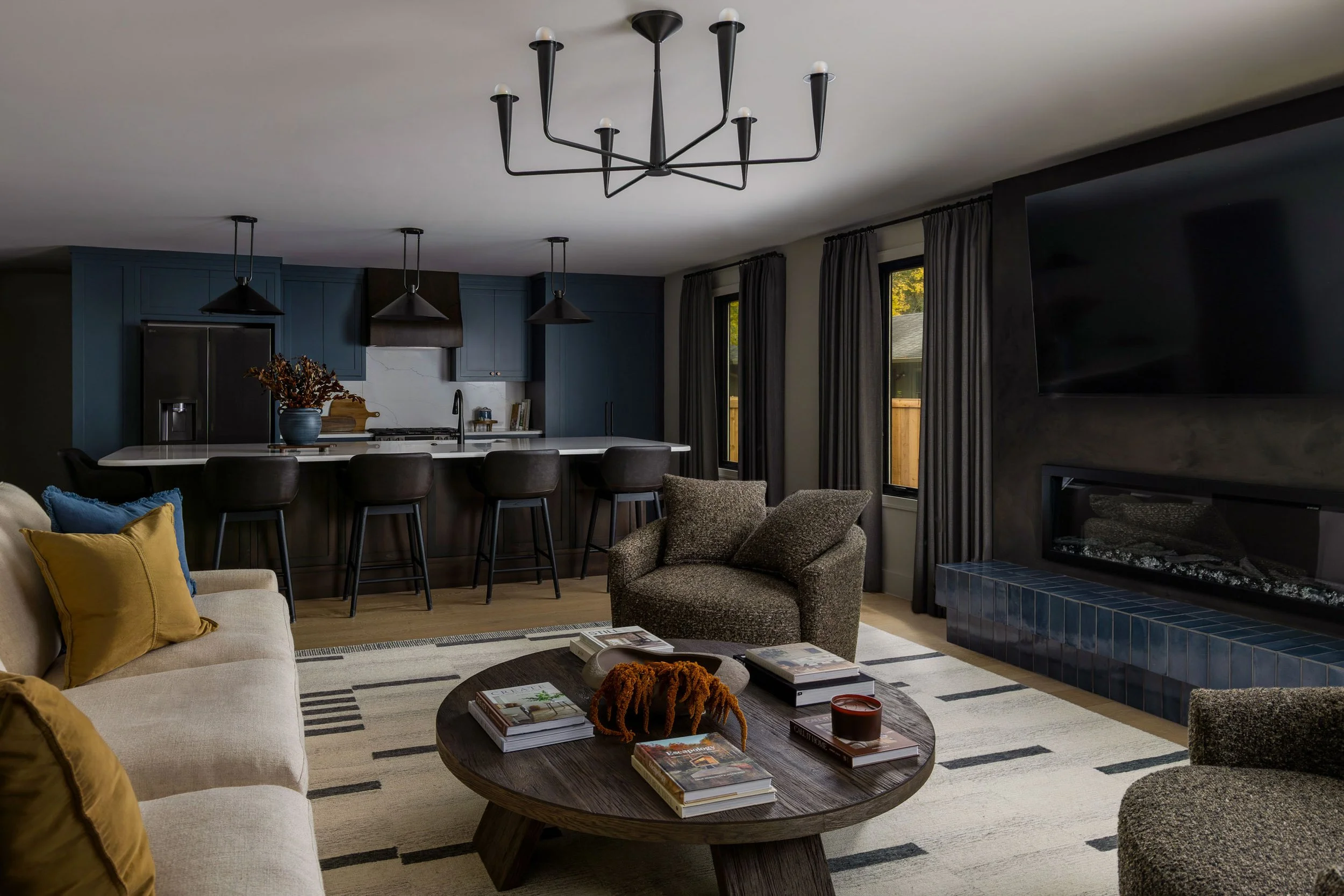 Living room with a white sofa, brown armchair, black coffee table with books and decorations, gray area rug, a fireplace with a blue tile surround, large windows with gray curtains, and a black chandelier.