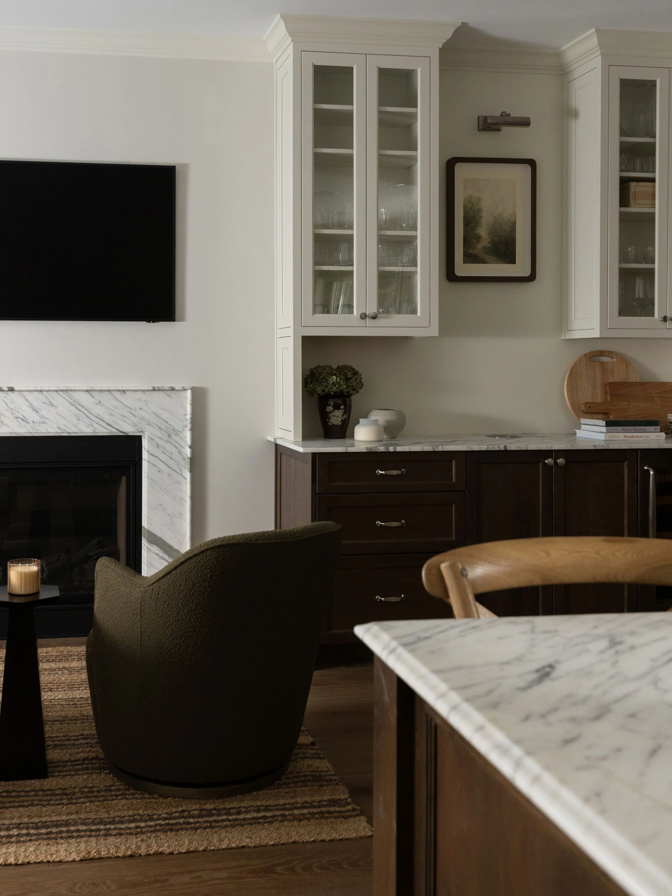 A living room with a flat-screen TV mounted on the wall, a fireplace with marble surround, a dark green armchair, a black side table with a candle, a patterned rug, white cabinetry with glass doors displaying glassware, a framed landscape painting, w