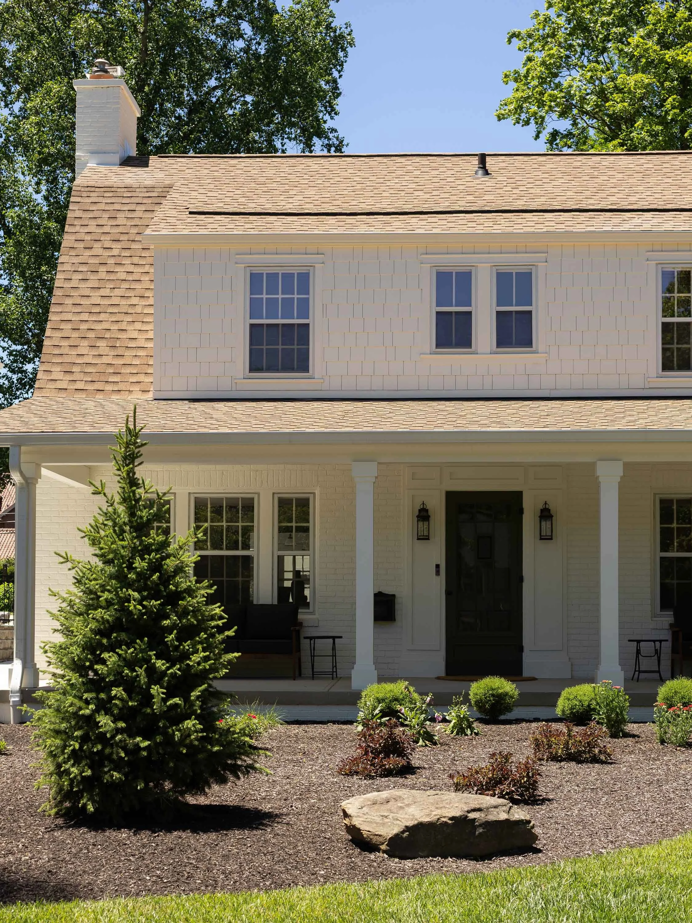 Front view of a white house with a porch, black door, and surrounding landscaped garden with bushes, a pine tree, and a large rock.