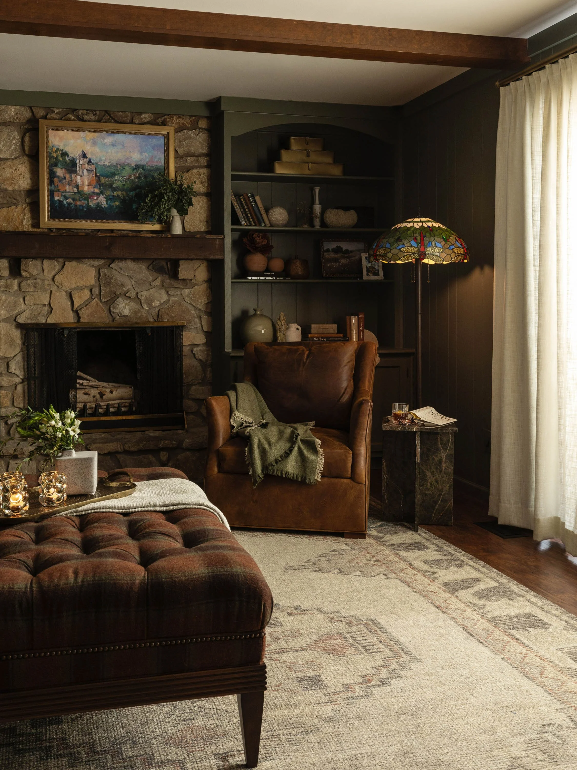 A cozy living room corner featuring a stone fireplace with a wooden mantle, a painting of a castle on a hill, a tall bookshelf with decorative items and books, a leather armchair with a throw blanket, a multicolored stained glass lamp, a side table w