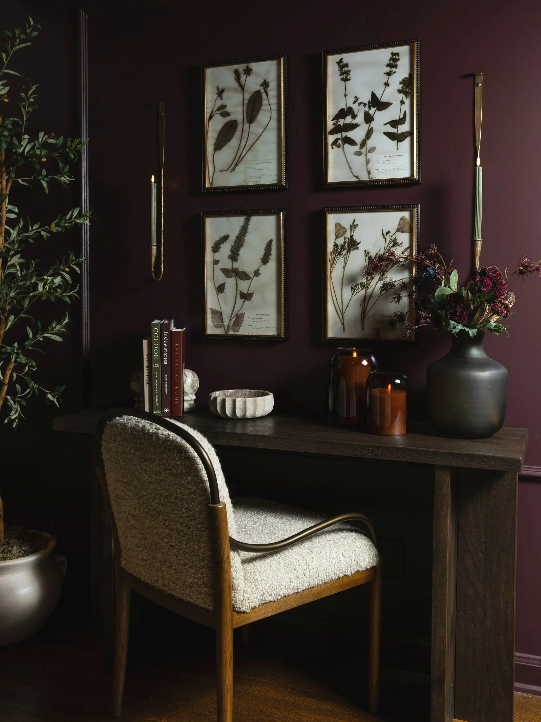 A cozy home workspace with a wooden desk, a cream-colored upholstered chair, and a dark maroon wall decorated with four framed botanical prints. There are potted plants, books, and candles on the desk.