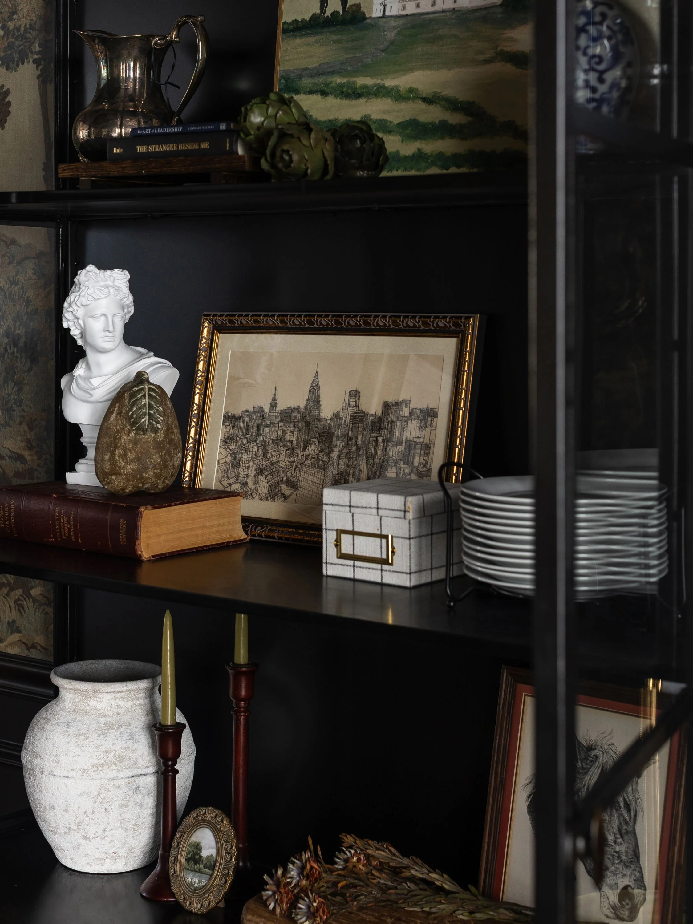 A black shelving unit displaying decorative objects, framed artwork, books, and ceramics. Items include a white bust sculpture, a large white textured vase, framed drawings of cityscapes, a silver pitcher, artichokes, and stack of white plates. There