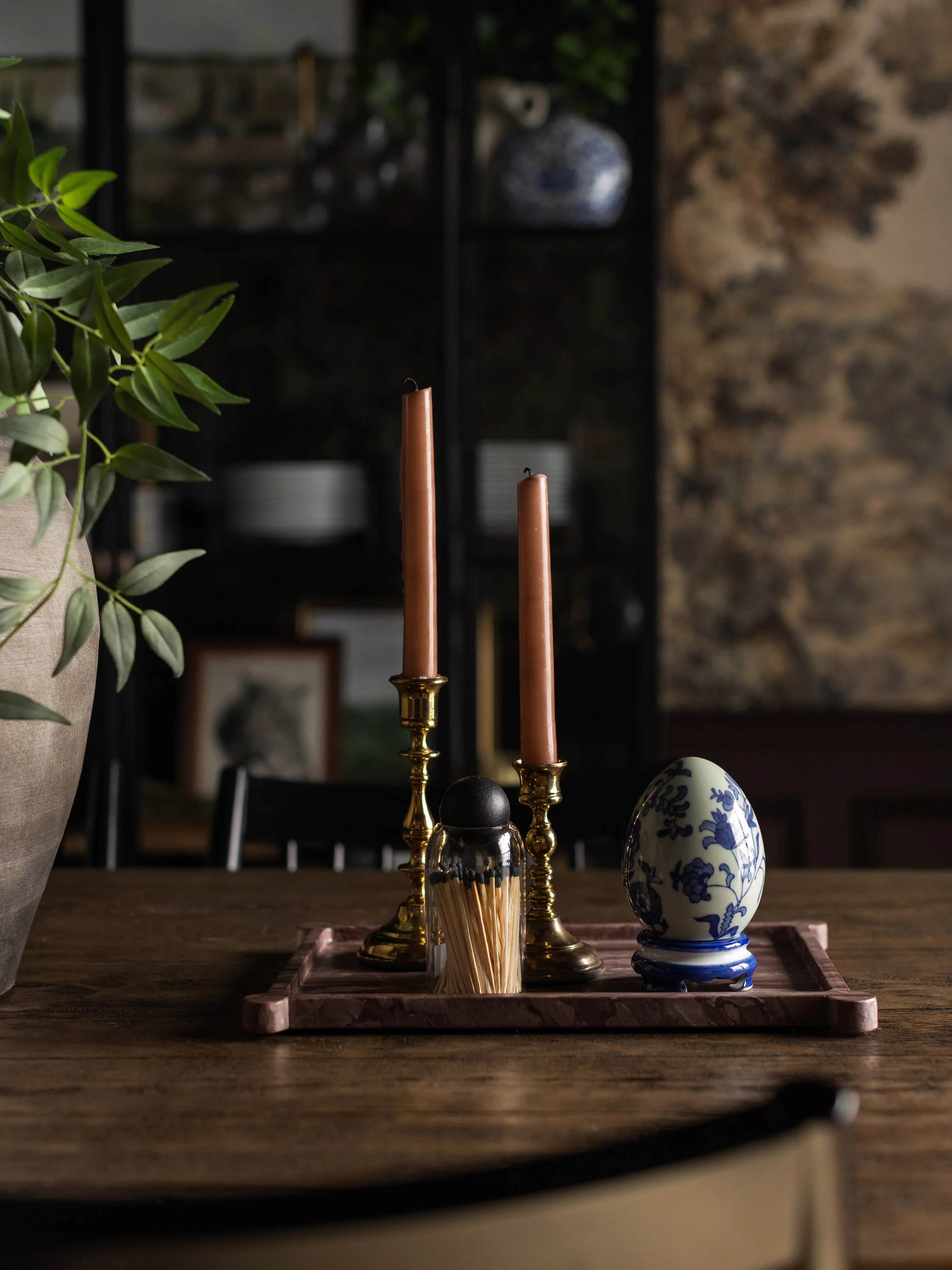 A wooden table with a tray holding two tall pink candles, a small jar with matches, and a blue and white ceramic egg-shaped ornament, with a plant on the left and a shelving unit in the background.