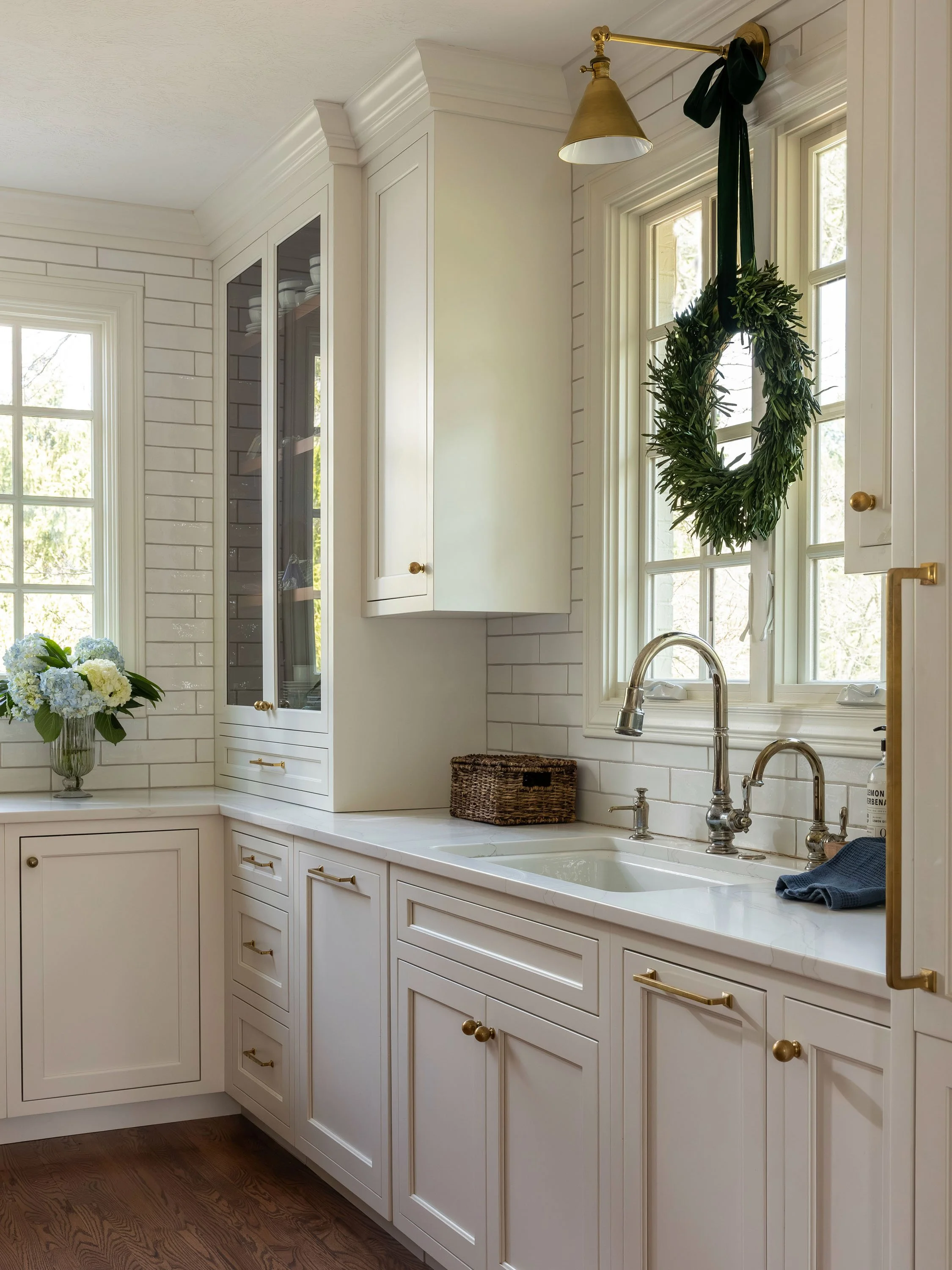 A kitchen with white cabinetry, a window with a wreath hanging on it, a faucet and sink, a basket, and a bouquet of flowers on the counter.