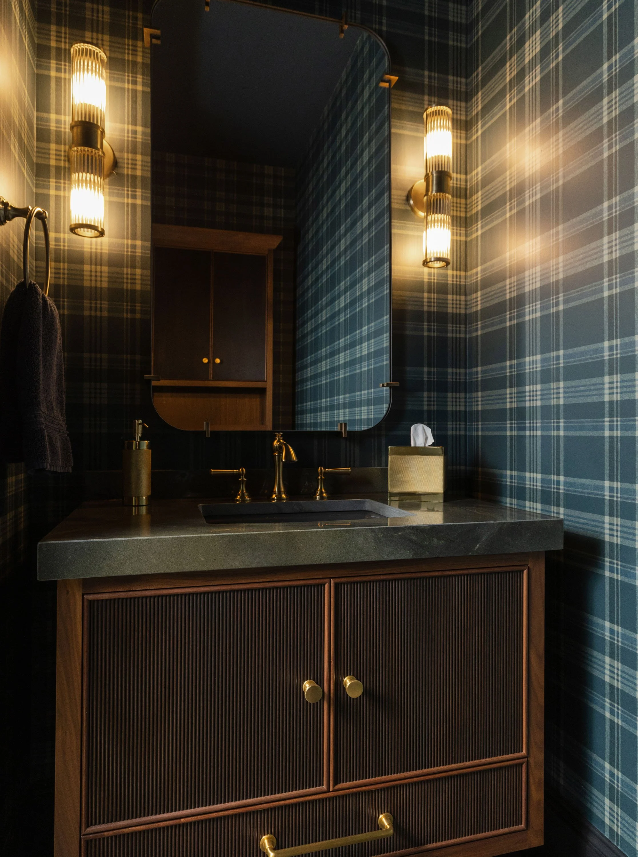A bathroom with plaid wallpaper, a mirror, and a wooden cabinet with golden knobs. There's a sink with a gold faucet, a soap dispenser, and a tissue box on the counter. Warm lighting is provided by wall-mounted lights.