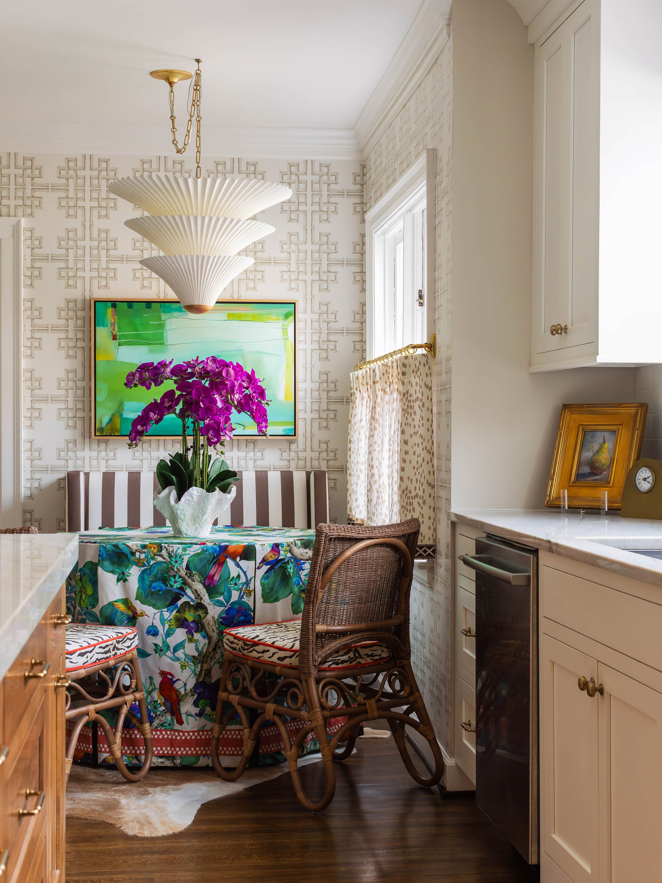 A kitchen with an intricate patterned wallpaper, a window with a leopard print curtain, a white cabinet, a bar fridge, a colorful tablecloth, a purple orchid in a white vase, a framed painting, a clock, and a hanging white ceiling light fixture.