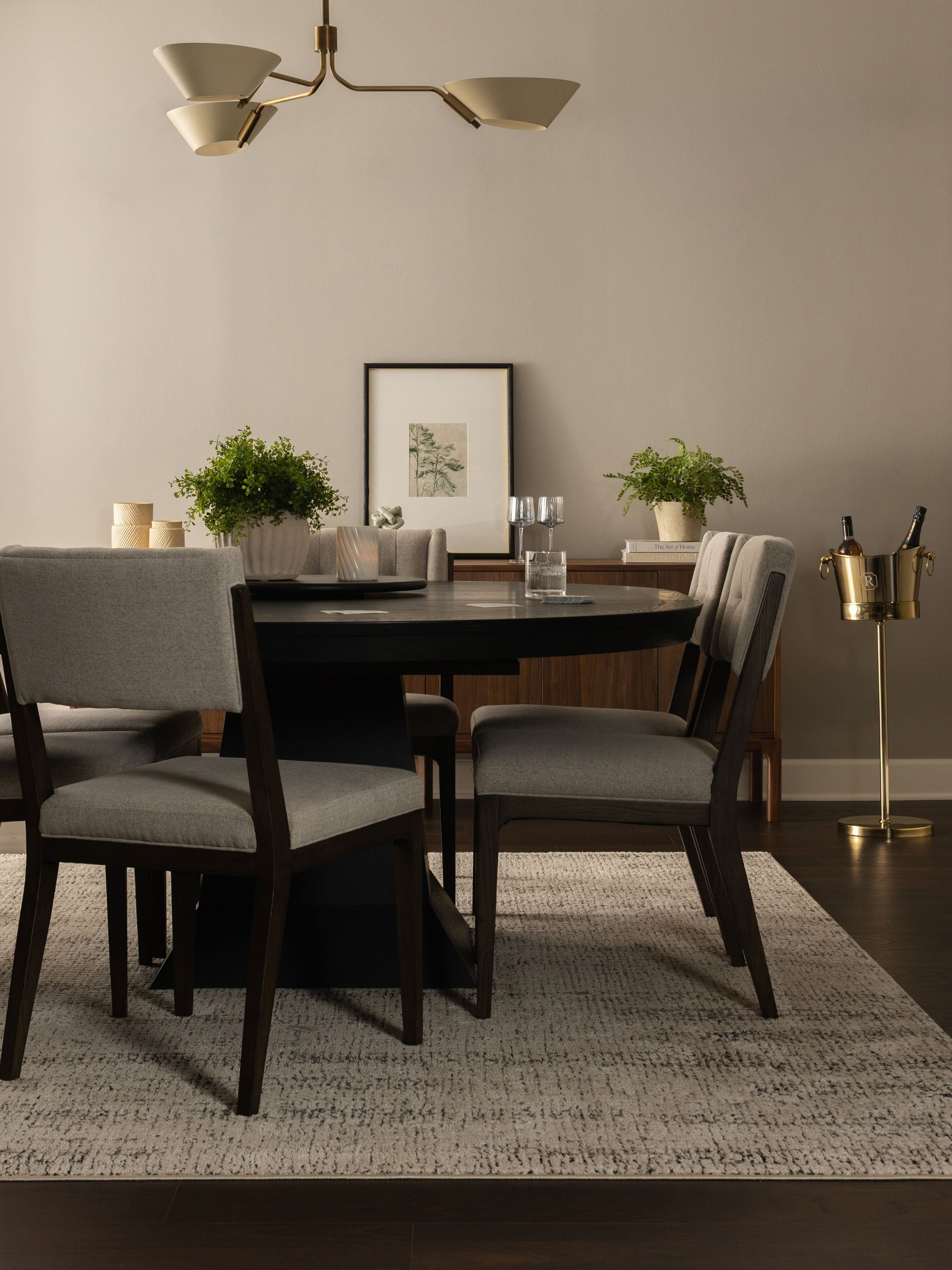 A modern dining room with a dark wood oval table, surrounded by upholstered chairs. There are two large potted plants on the table, a framed artwork on the wall behind, and a sideboard with books and decorative items. A gold floor lamp is to the righ