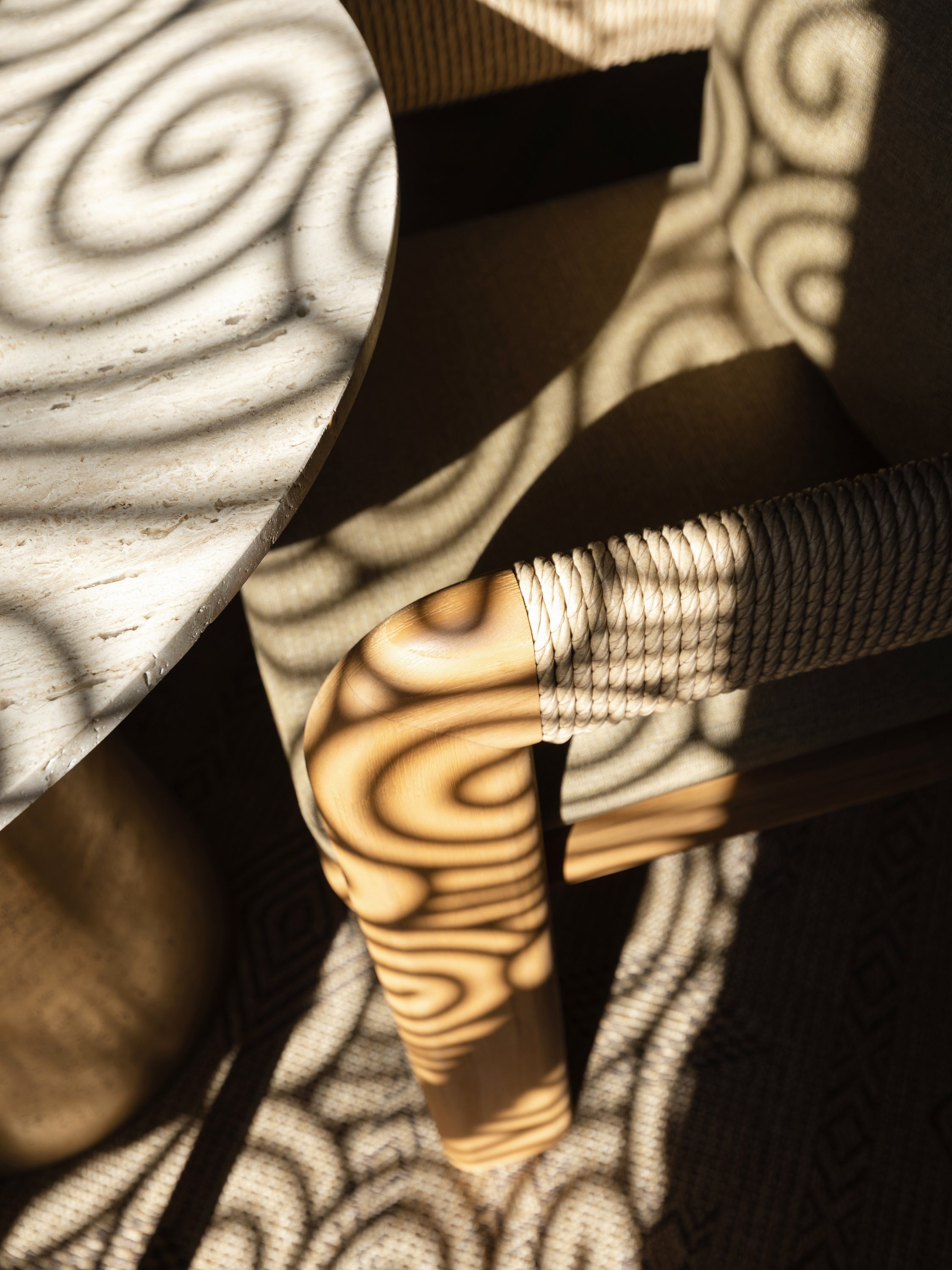 Close-up of a chair with a woven rope armrest and a wooden leg, casting shadows on a fabric surface.