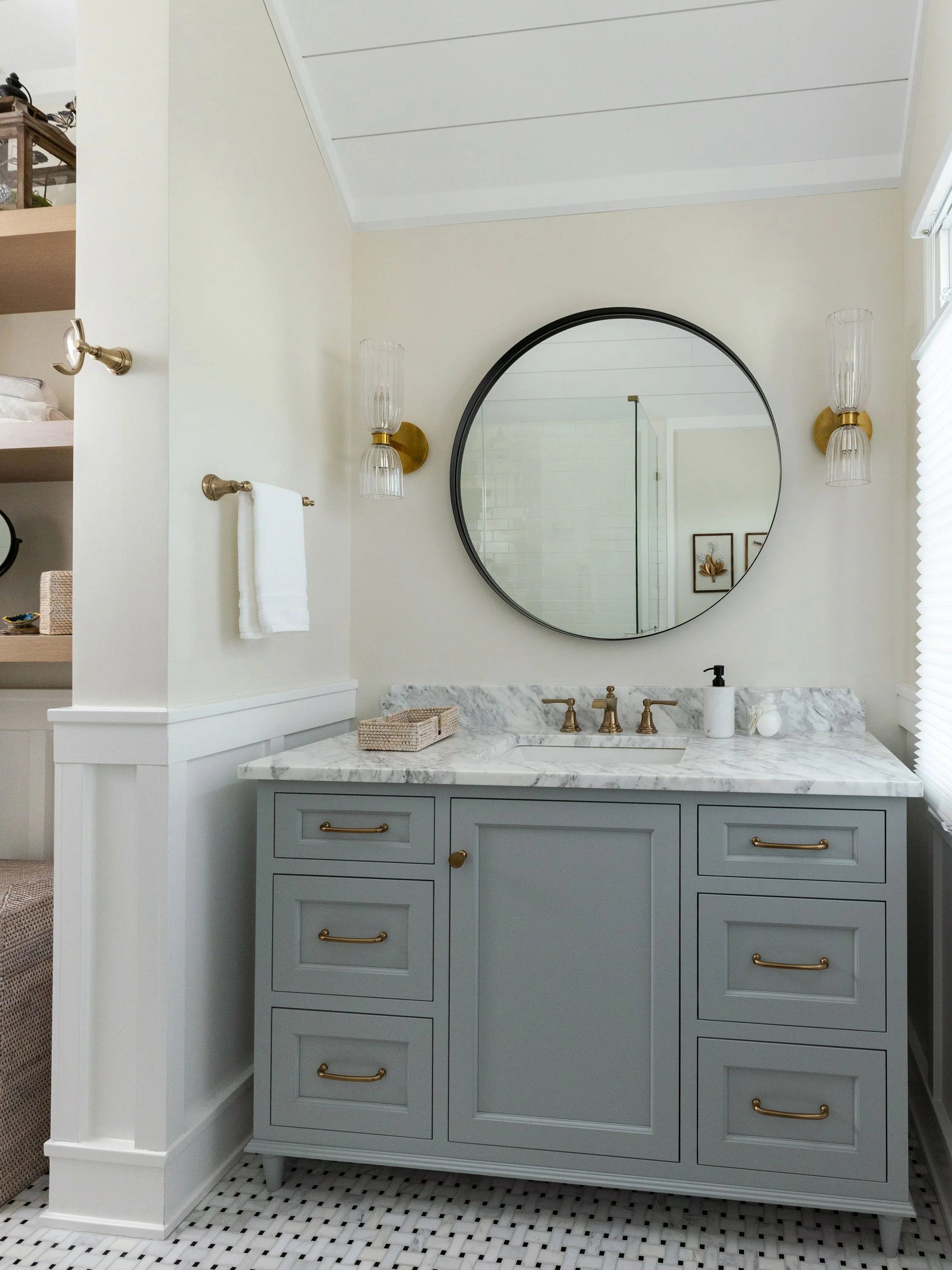 Bathroom vanity with a gray cabinet, marble countertop, and gold hardware. A round mirror and two wall-mounted glass light fixtures are above the sink. The floor has small black and white tiles. There is a window with blinds on the right.