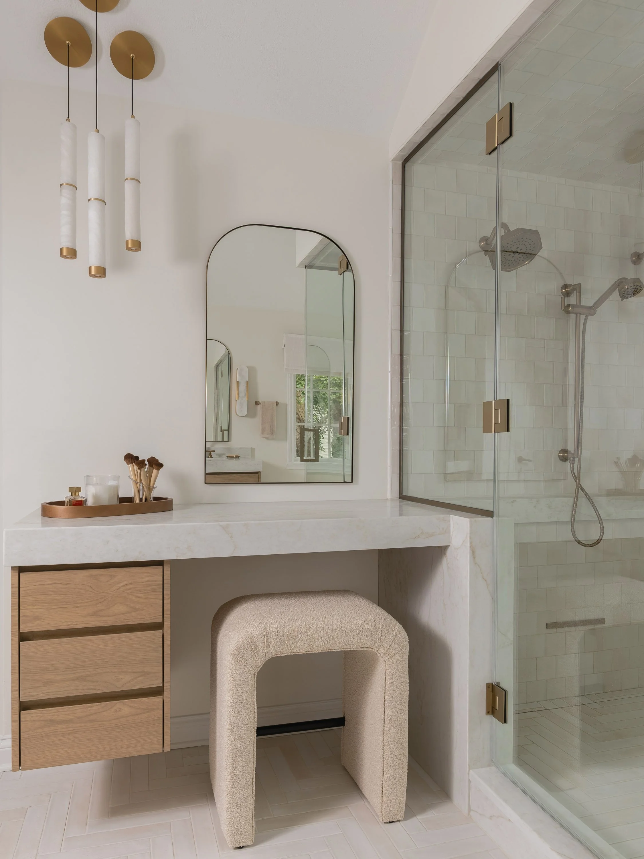 Modern bathroom with a marble vanity, a large mirror, a beige upholstered stool, a small vanity table with makeup brushes and skincare products, and a glass-enclosed shower with white subway tiles.
