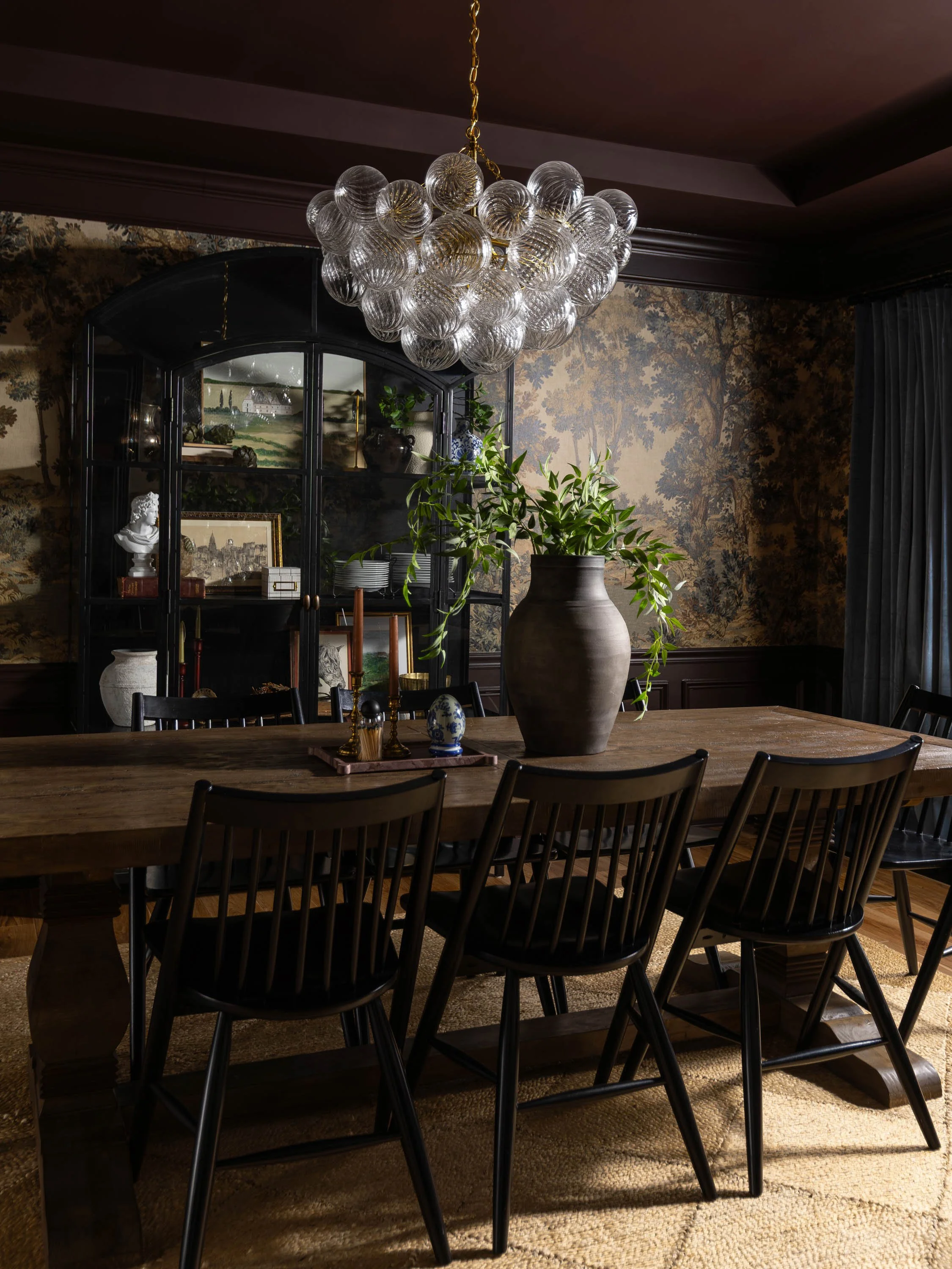 A dining room with a large wooden table surrounded by black chairs, a large vase with greenery on the table, a black display cabinet with decorative items and framed artwork inside, a chandelier with glass globes hanging from the ceiling, patterned w