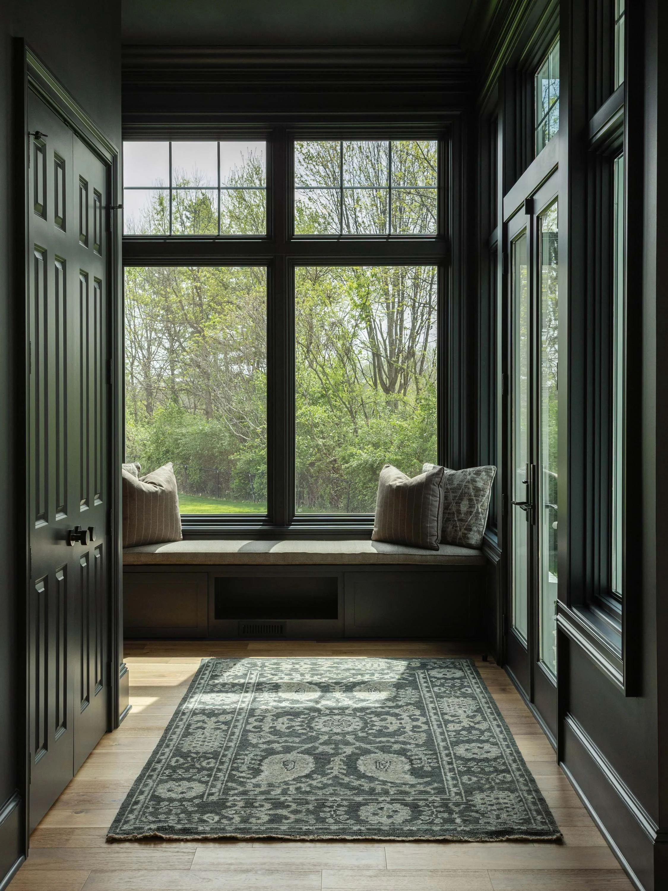 View of a cozy bay window nook with a cushioned window seat and pillows, overlooking a lush green outdoor scene, with black window frames and trim, and a patterned rug on wooden flooring.