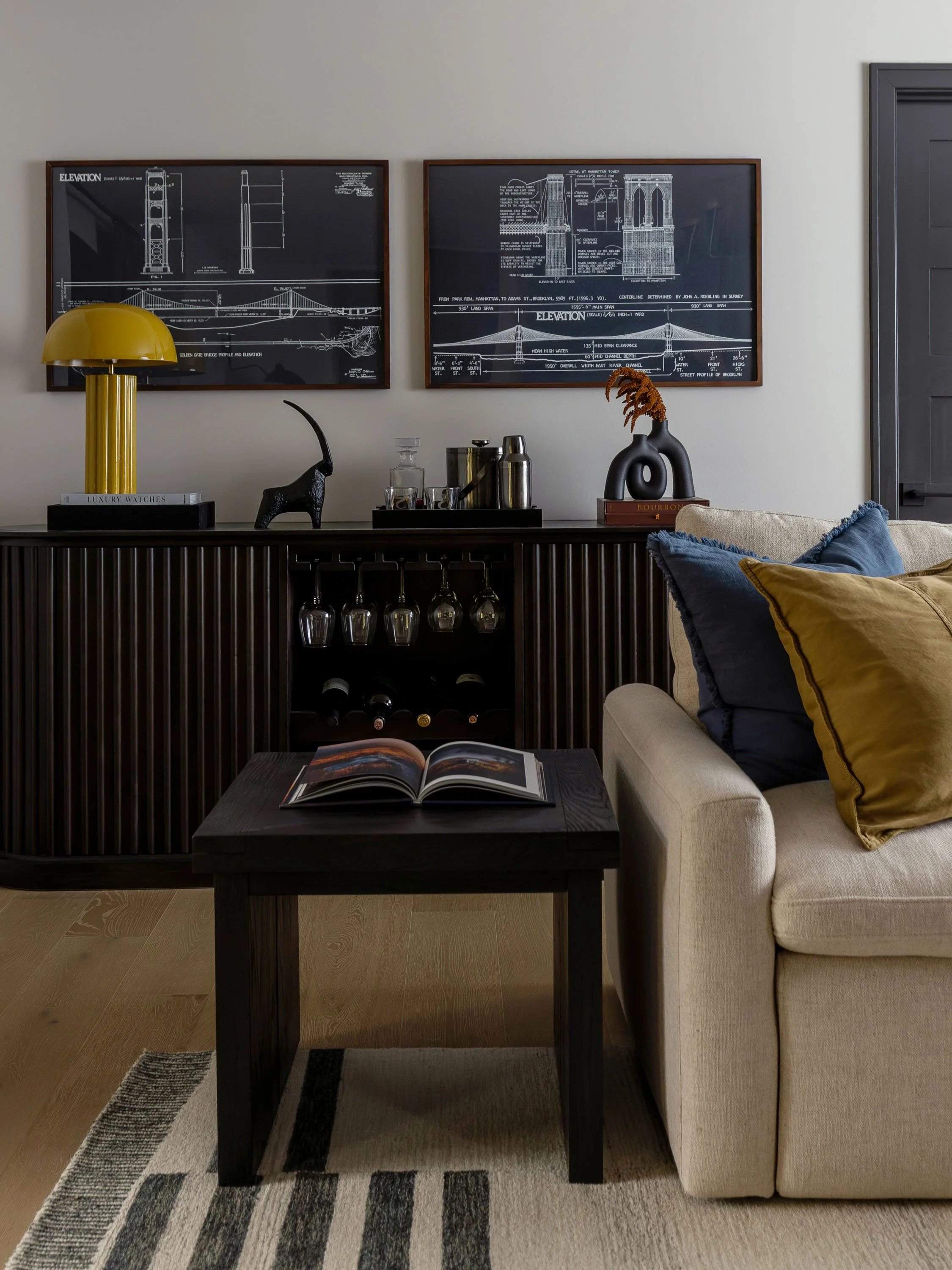 A living room with a beige sofa with yellow and dark blue pillows, a black coffee table with an open book, a black sideboard with glassware, a yellow modern lamp, and two framed architectural blueprints on the wall