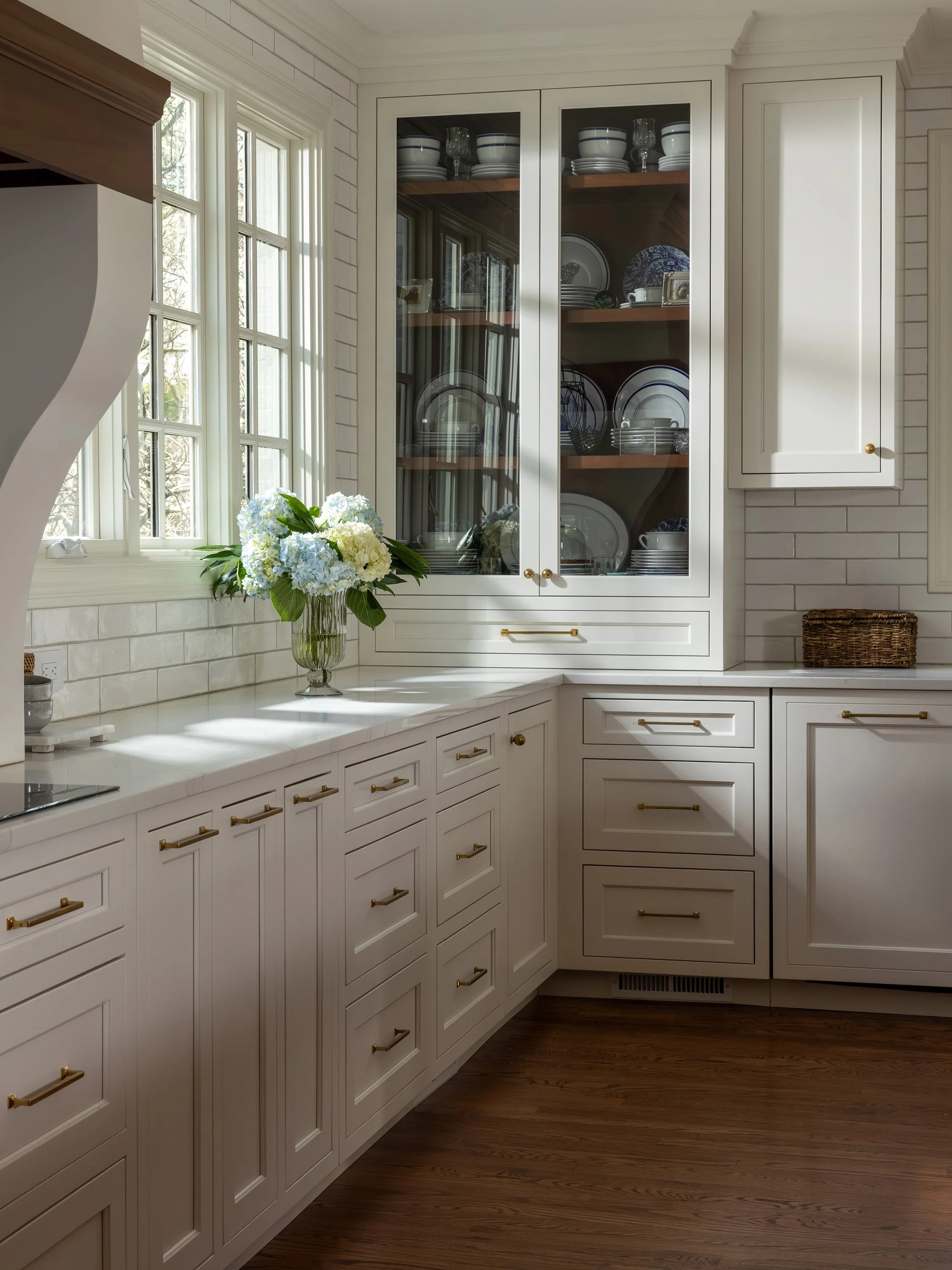 A corner of a kitchen with white cabinetry, gold handles, a white tiled backsplash, a window, a vase of white and blue flowers, and a woven basket.