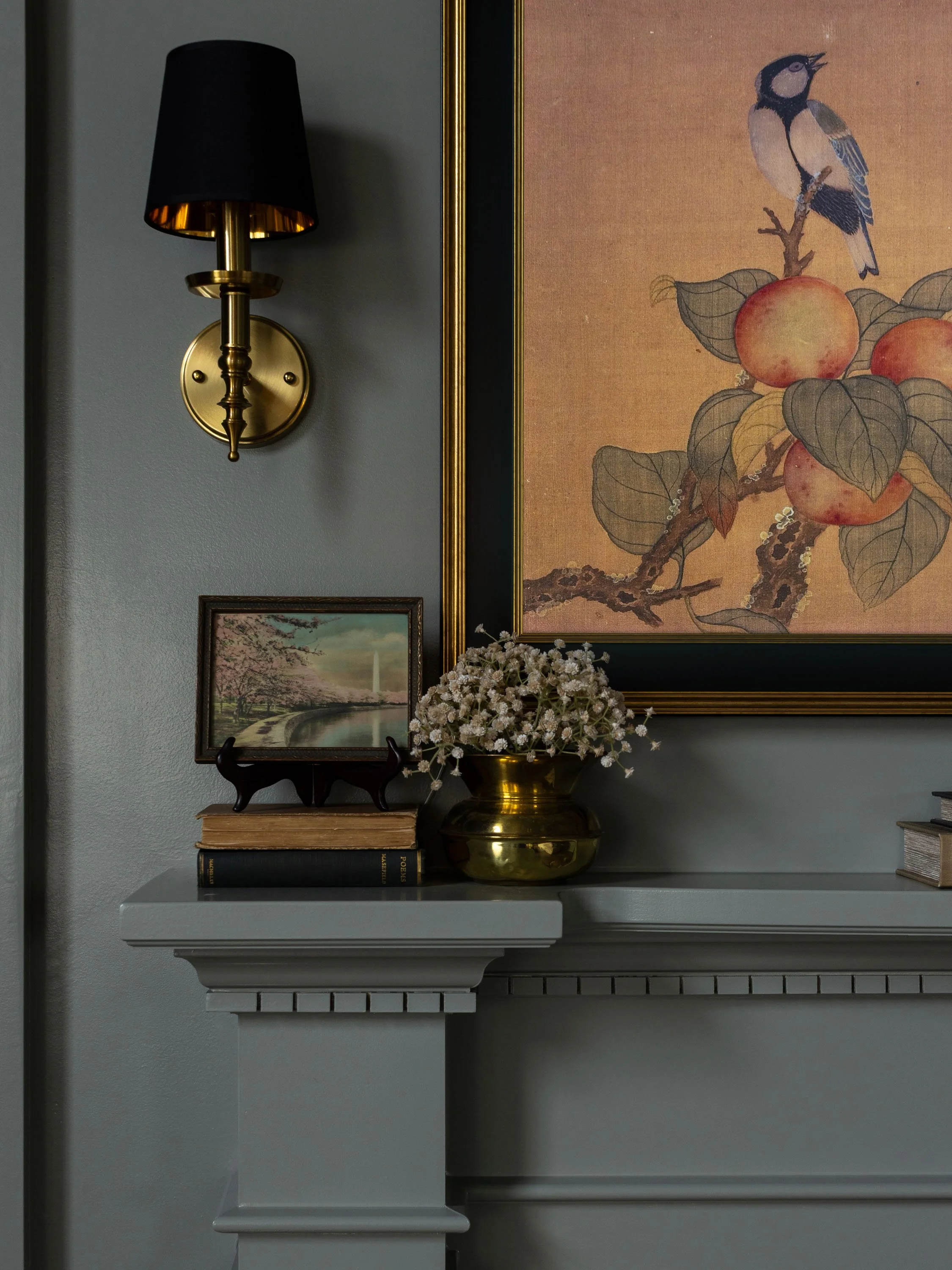 A living room wall decorated with a black and gold wall sconce, framed artwork of a bird sitting on a branch with peaches, a landscape photo of cherry blossoms and a fountain, and a white mantel with a gold vase of white flowers and books.