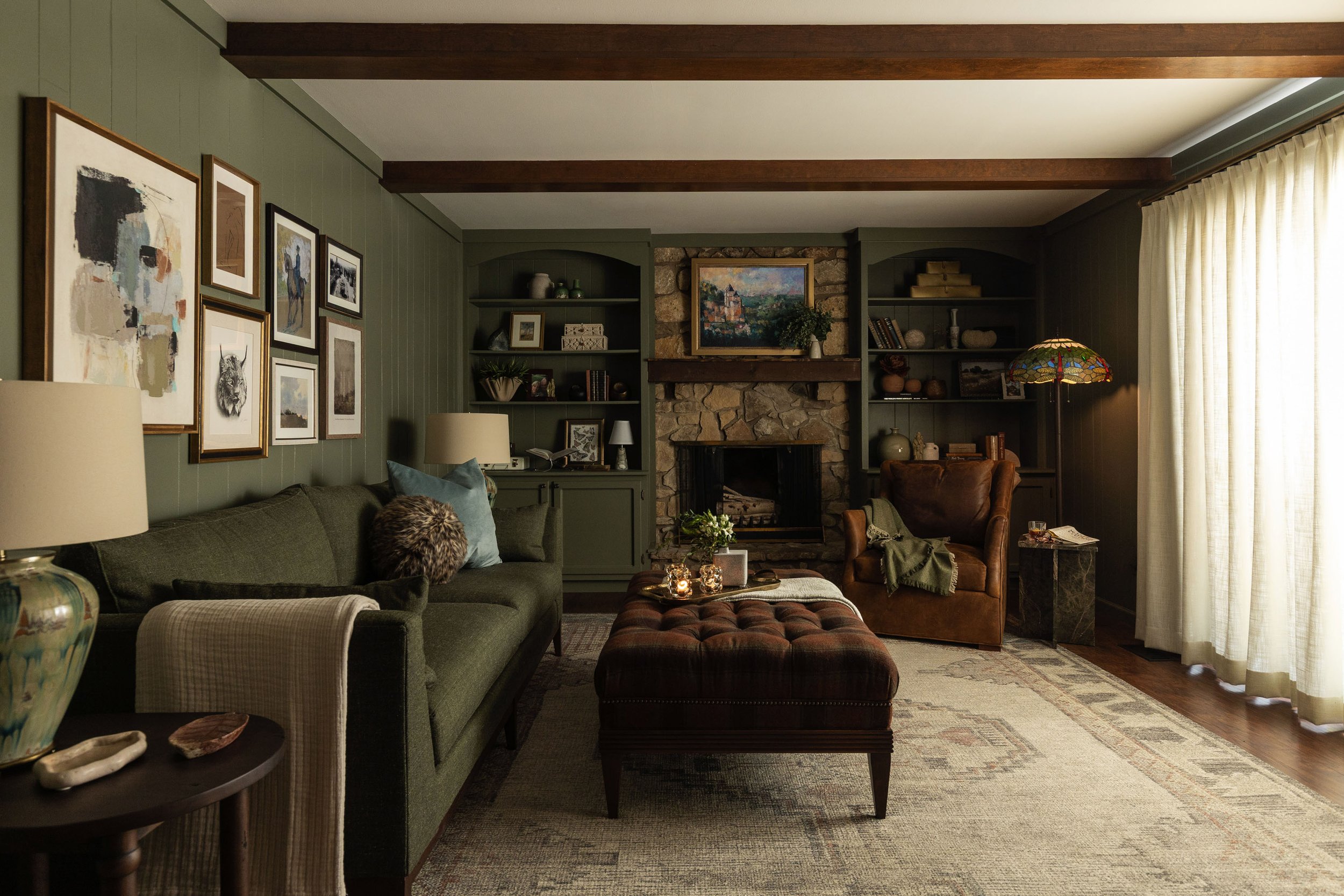 A cozy living room with a green couch, a leather armchair, and a patterned ottoman. There are various paintings and photographs on the green walls and a stone fireplace with a framed landscape painting above it. A bookshelf with decorative items, candles, and books is on the right side. A floor lamp with a colorful stained-glass shade, a table with candles, and beige curtains covering large windows are also visible.