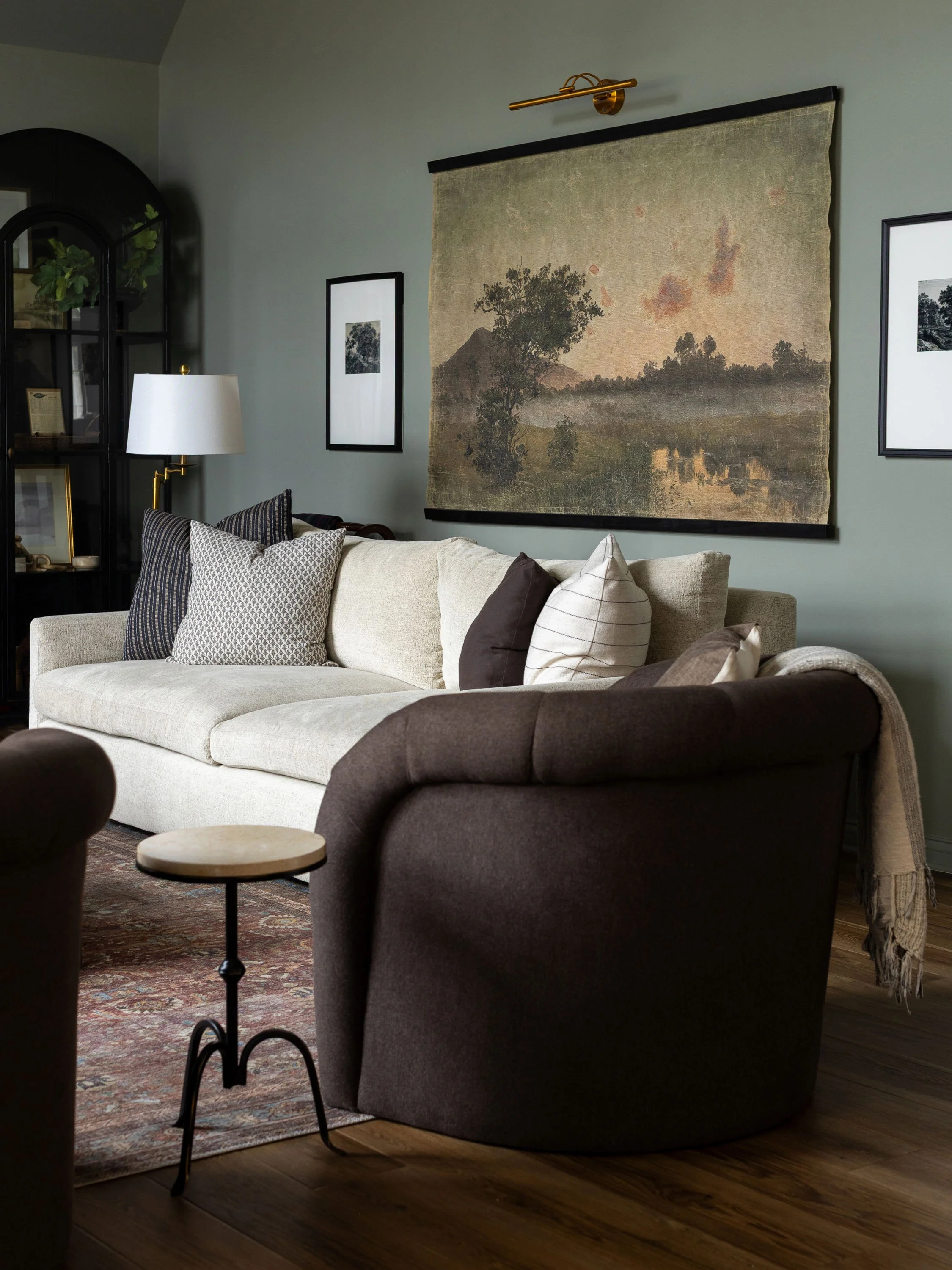Living room with a beige sofa, dark armchair, various pillows, a side table, a patterned rug, a wall tapestry depicting a landscape, framed pictures, a white lamp, and a black display cabinet with decorative items.