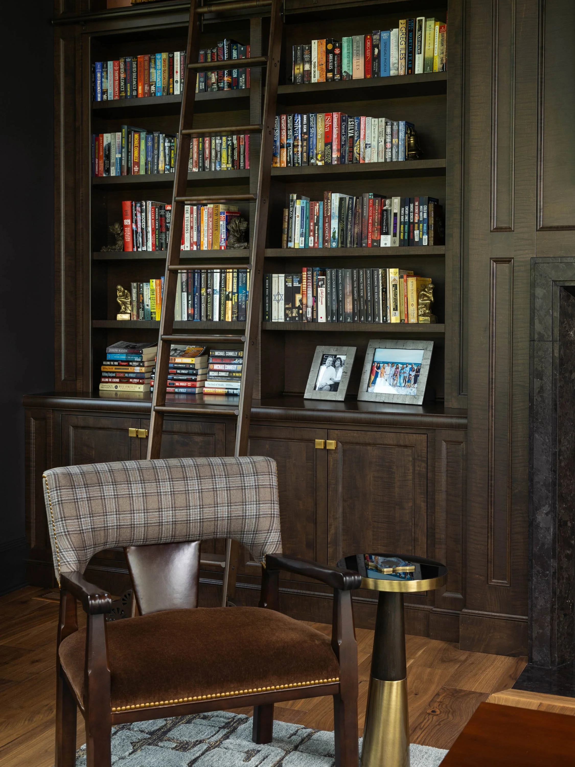 A cozy reading or sitting area with a wooden bookshelf filled with books, a ladder for reaching high shelves, a plaid vintage-style armchair, a small side table with a gold-colored base, and framed photographs on the bookshelf against a dark wood-pan