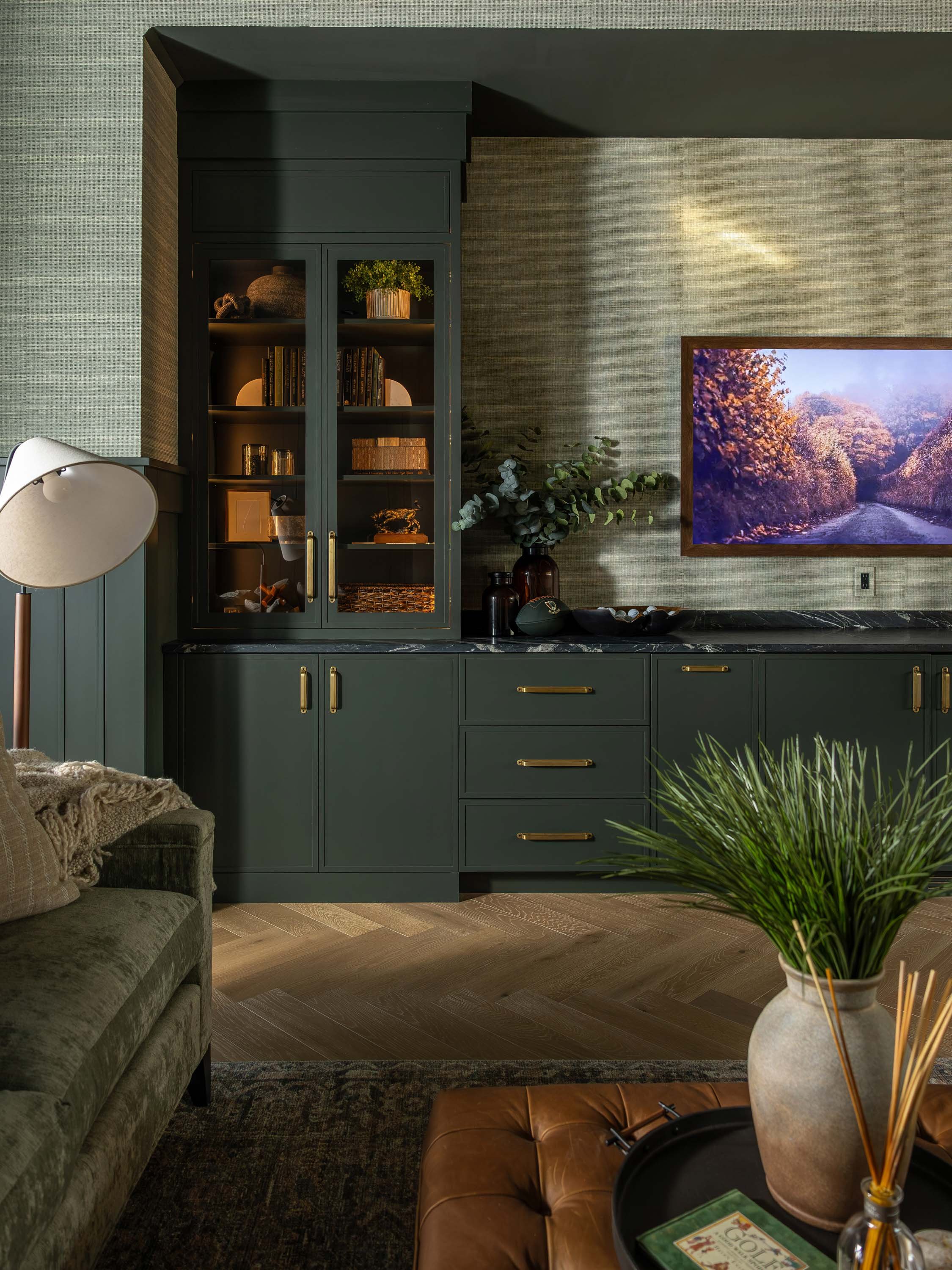 Living room with a dark green cabinet, a green sofa with pillows, and a round table with a vase, magazines, and candles, featuring hardwood floors and textured wallpaper.