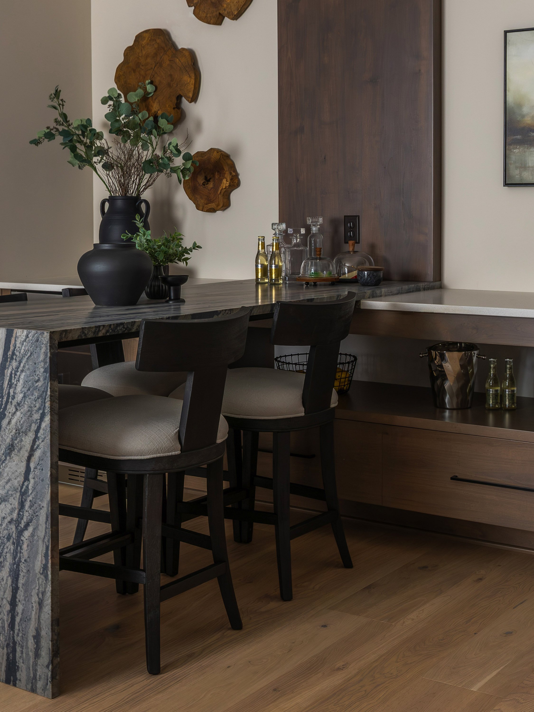 Interior view of a dining area with a wooden table, black chairs with beige cushions, decorative green plants in black vases, and a wall with wooden art pieces. Some bottles and glassware are on the table, and there is a power outlet on the wall.