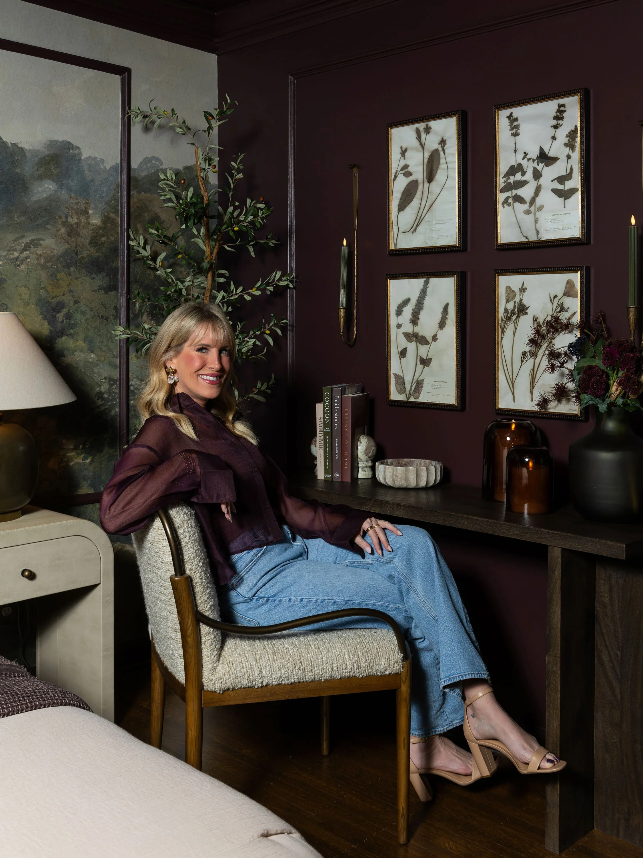 A woman sitting in a vintage armchair in a cozy, decorated room with dark walls, framed botanical prints, and various decorative items on a table.