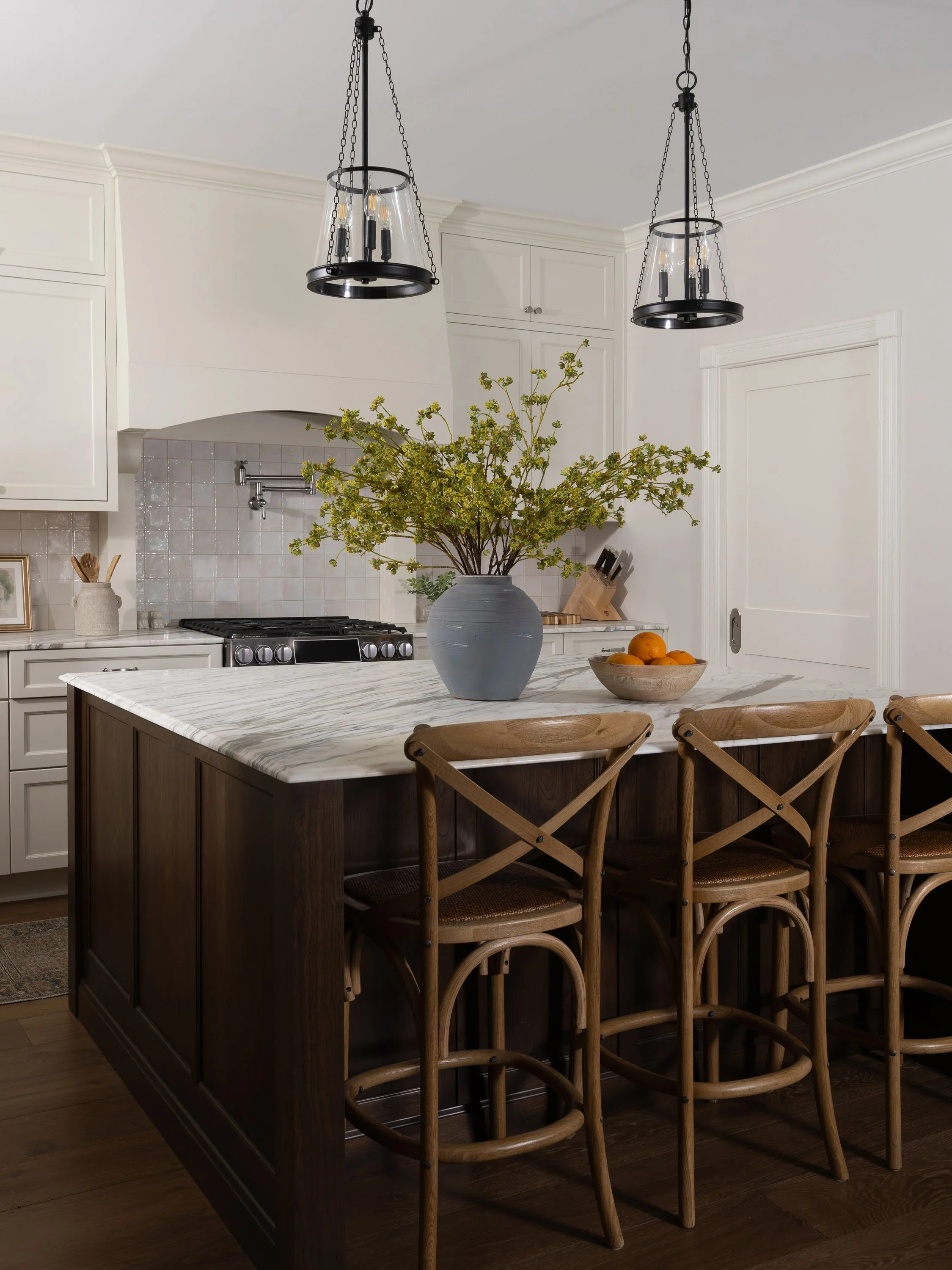The image shows a modern kitchen with a large island with a marble countertop, surrounded by four wooden chairs. There is a blue vase with green branches and a bowl of oranges on the island. Two black pendant lights hang from the ceiling. The backgro