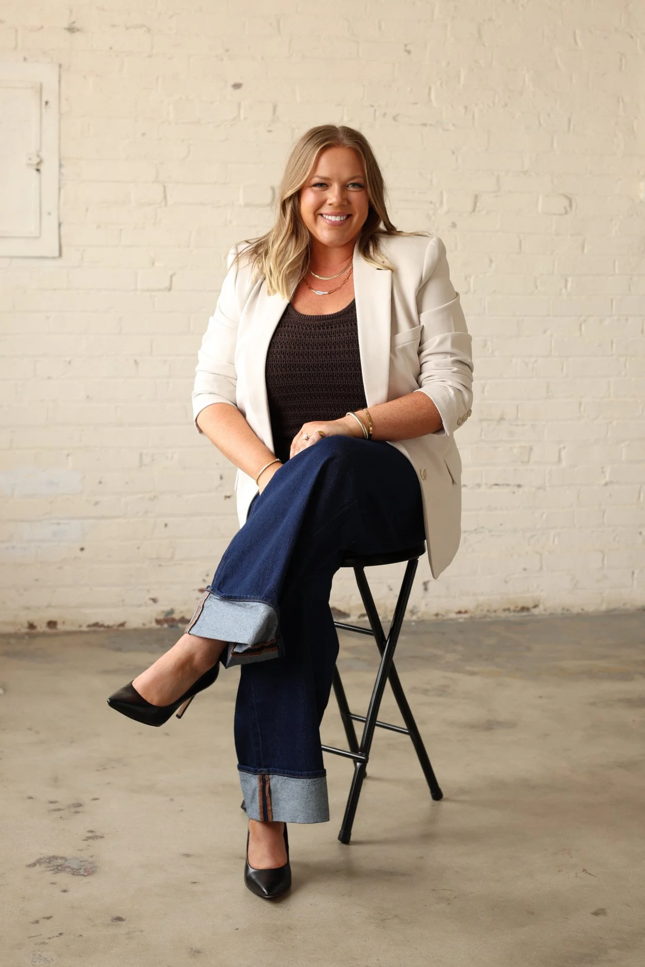 A woman with blonde hair smiling, seated on a black metal chair, wearing a white blazer, black top, blue jeans with cuffed hems, black high heels, and gold jewelry, sitting against a white brick wall in an industrial-style setting.