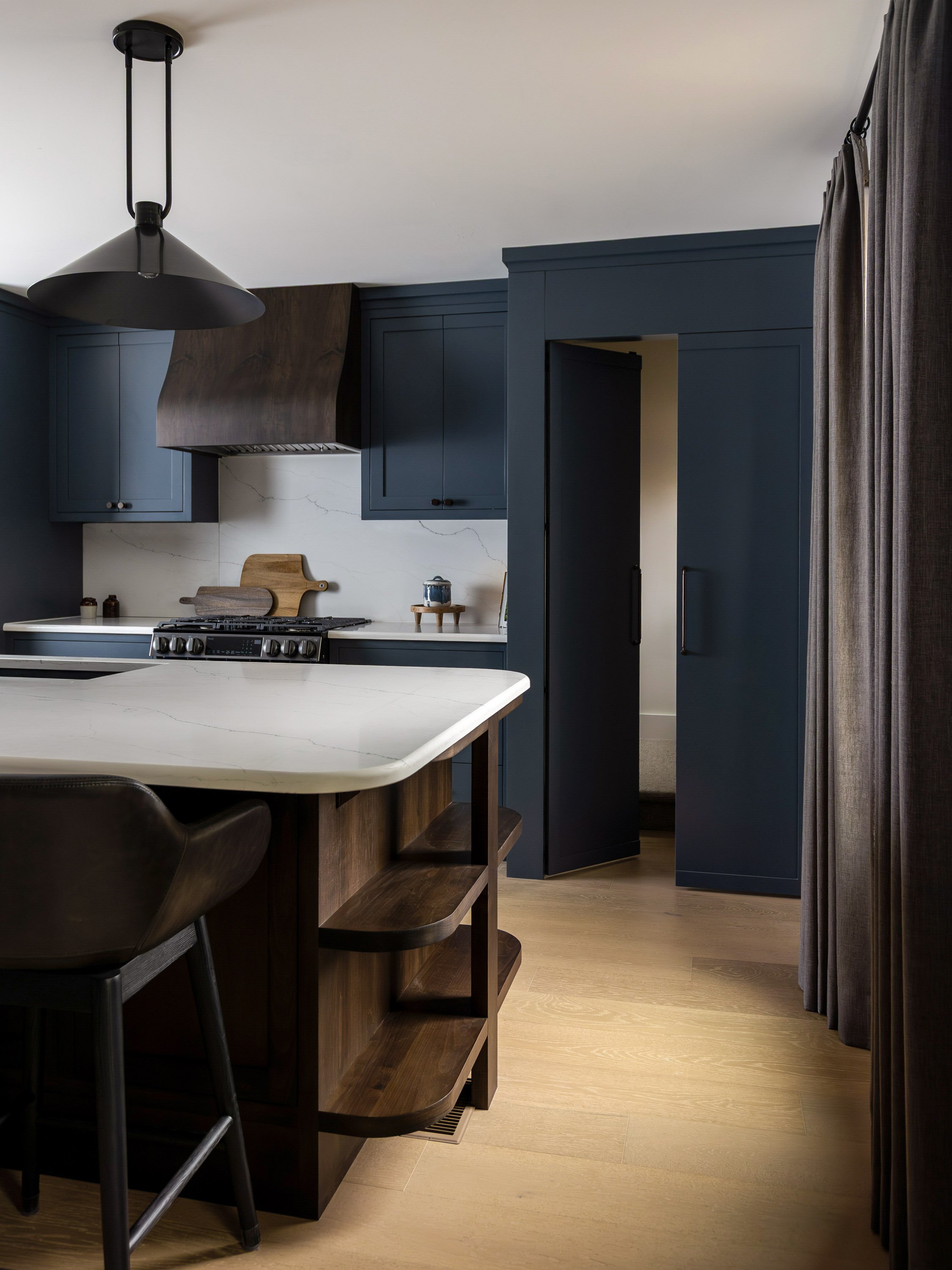 Interior view of a modern kitchen with blue cabinets, a wooden kitchen island with bar stools, and a white marble countertop. There is a black pendant light hanging from the ceiling and a gray curtain on the right side.
