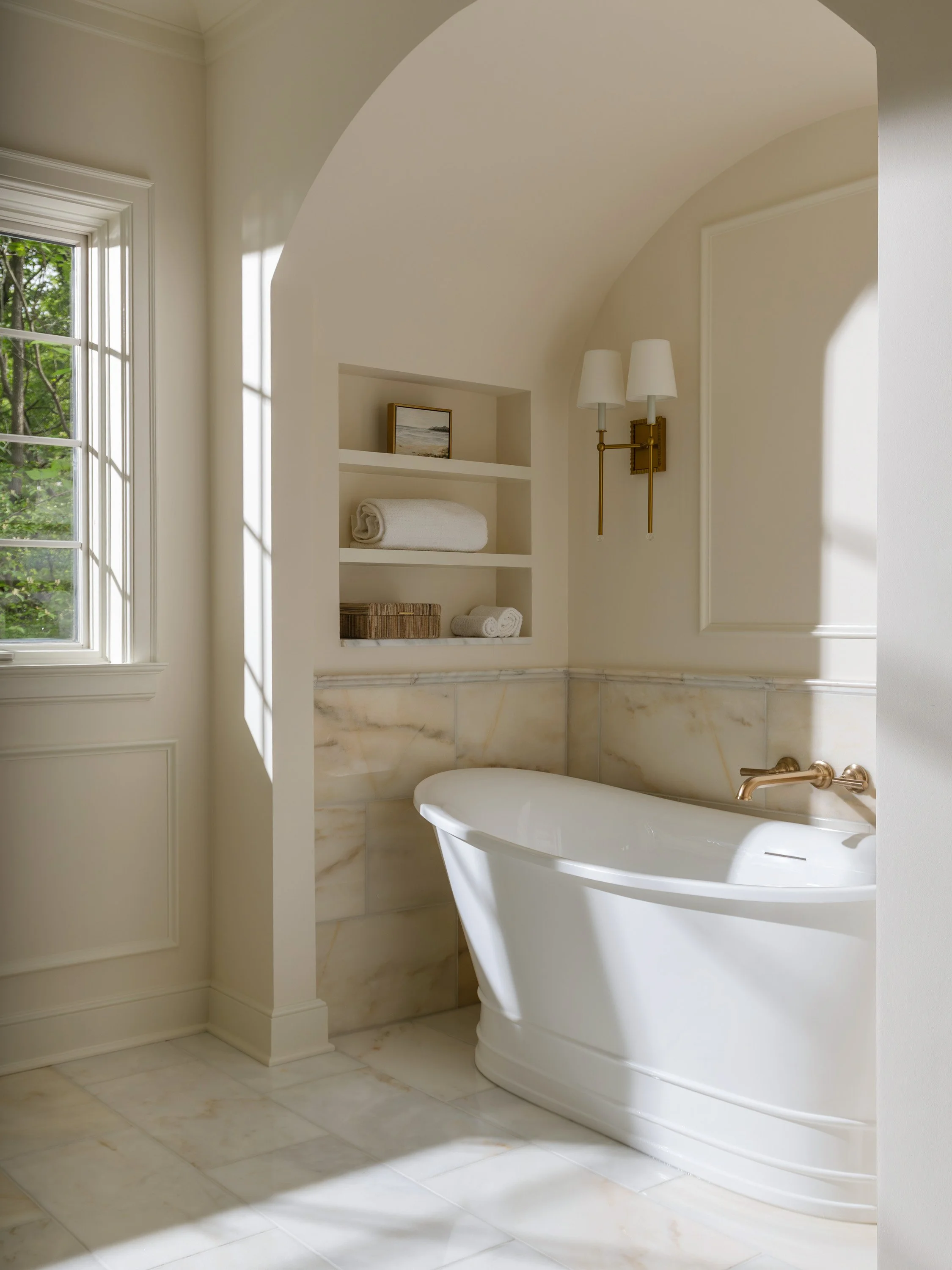 A white bathtub in a bathroom with a marble floor and walls, a window letting in natural light, open shelving with towels and a small framed picture, and a wall-mounted light fixture.