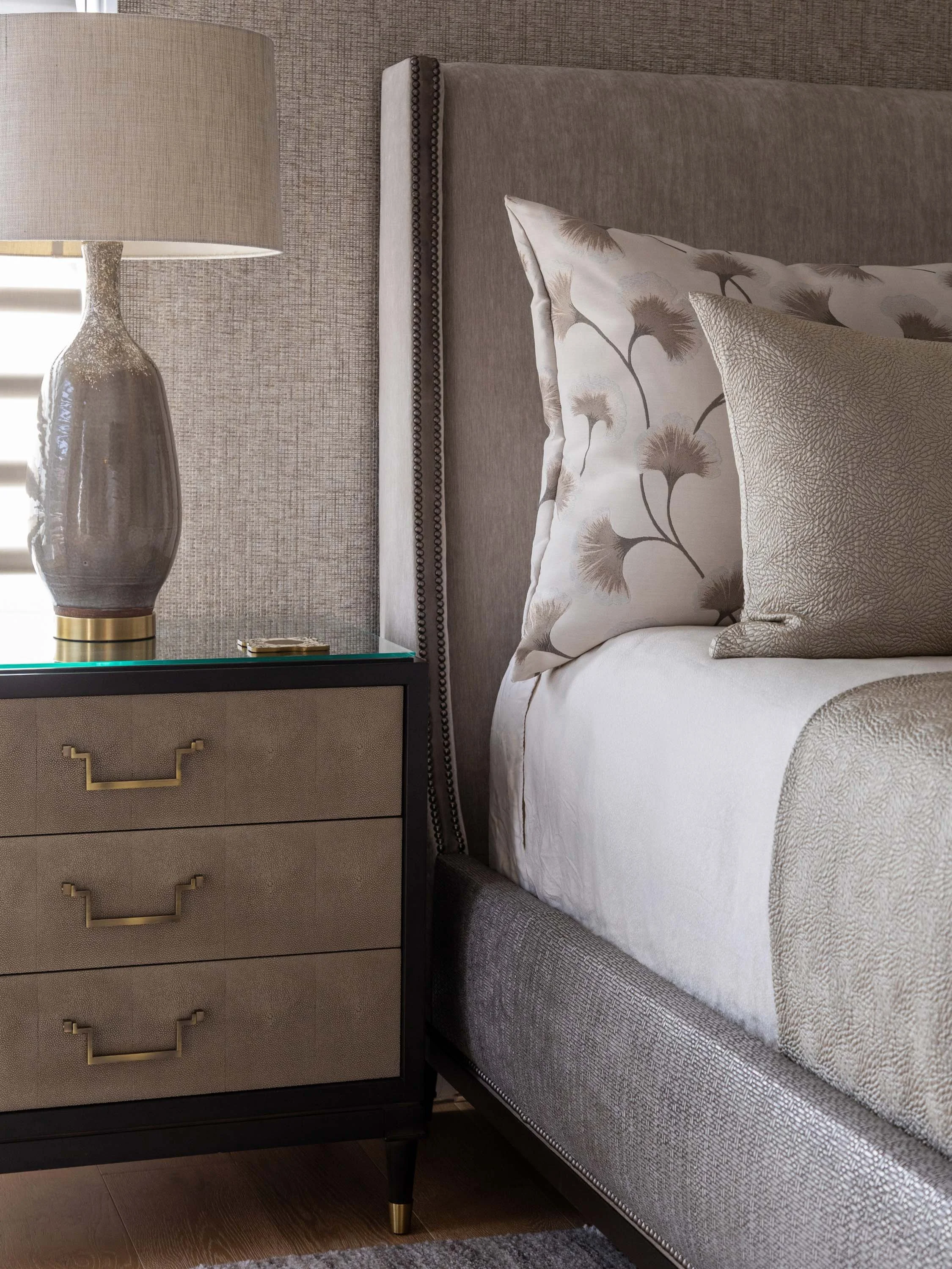 Close-up of a bedside table with a lamp and decorative objects next to a bed with pillows, a tufted headboard, and bedding in neutral tones.